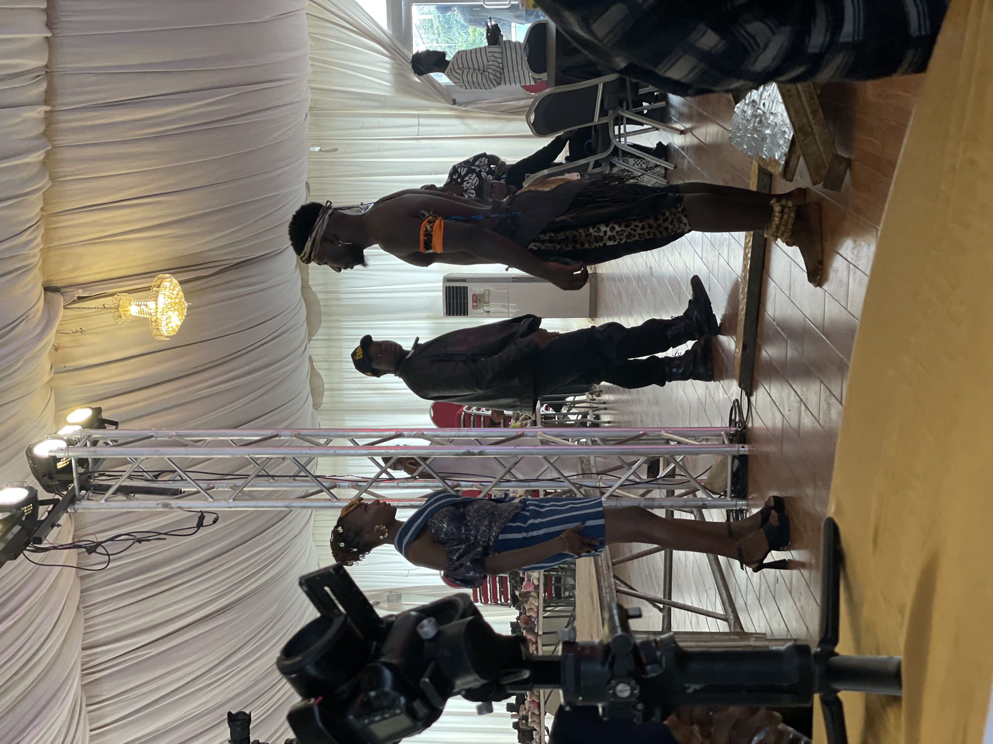 This media file captures an indoor event in Barakin Lerawi, Nigeria, likely a celebration or performance. The scene is set within a draped tent, suggested by the abundance of white fabric creating a soft, diffused background. A prominent crystal chandelier hangs from the ceiling, casting a warm glow and indicating an evening or dimly lit indoor setting.
The primary subjects are several individuals, seemingly engaged in a presentation or performance. In the foreground, a person in traditional attire, adorned with a patterned skirt and beaded anklets, stands looking upwards. Behind them, a person in a black leather jacket and cap is also present, standing stoically. Further back, another individual, shirtless and wearing a headband and an orange armband, is positioned looking down. In the background, a person in a striped shirt is seated, looking at a phone.
The event appears to involve some form of performance or ritual, given the formal attire of some individuals and the stage-like setup with lighting rigs and truss structures. The lighting suggests it's an indoor event, possibly in the evening. The floor is tiled with a warm wooden pattern. The overall mood is one of anticipation or engagement with the ongoing activity. No visible text is present in the media file.