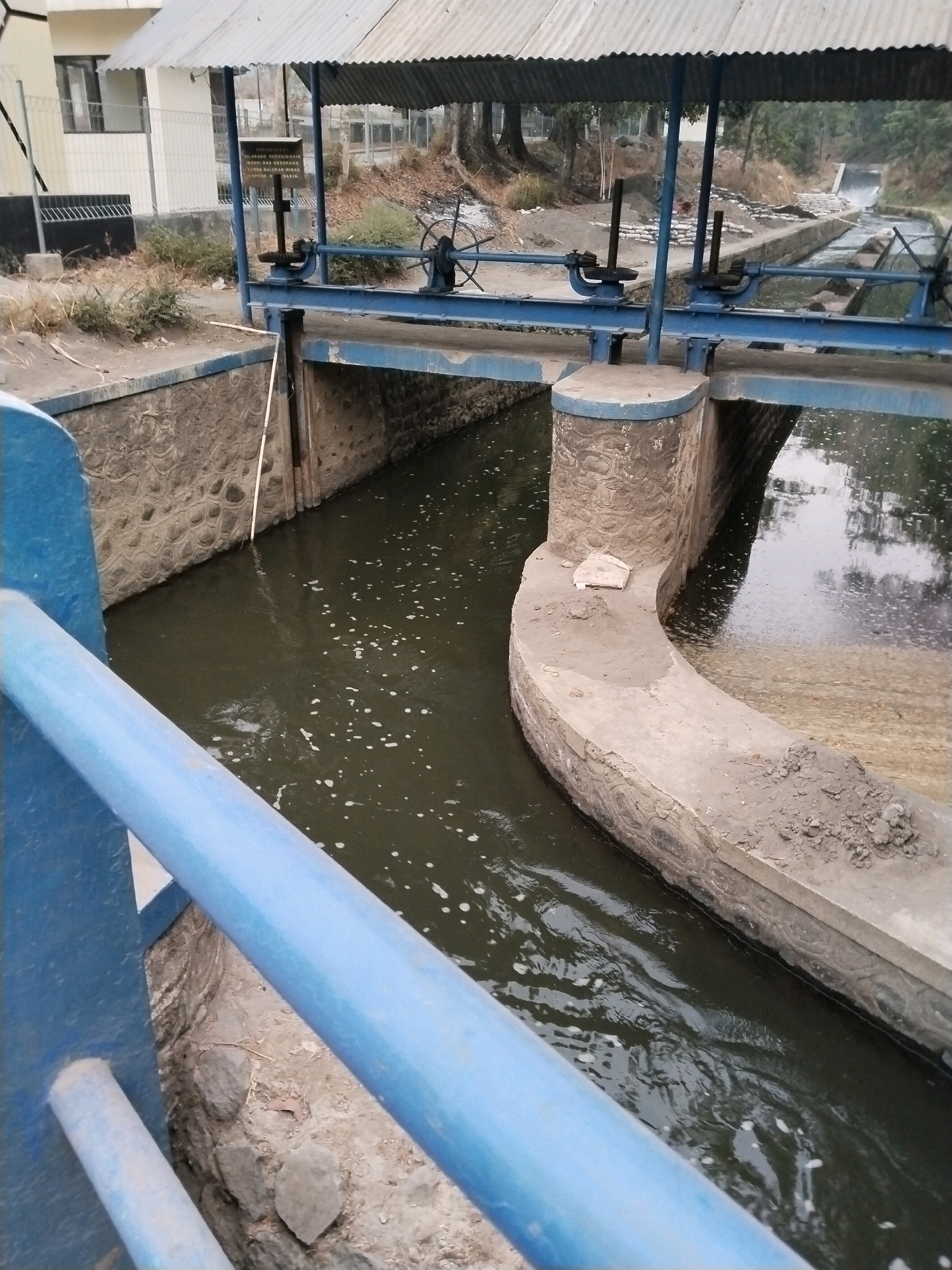 The image captures a section of a water control structure, likely a weir or dam, located along a channel in Sangkanjaya, Indonesia. The structure features a metallic framework painted blue, supporting a corrugated metal roof. Two manually operated gates, each with a wheel mechanism for adjustment, are visible. The water in the channel appears dark and somewhat murky, with some floating debris or foam. The channel walls are constructed from stone and concrete. Beyond the immediate structure, a more distant view shows a stepped waterfall in a different part of the waterway, suggesting a multi-level water management system. The surrounding environment includes vegetation, dry earth, and buildings in the background, indicating an outdoor, possibly industrial or utility setting. The perspective is from a raised vantage point, looking down and across the water channel.