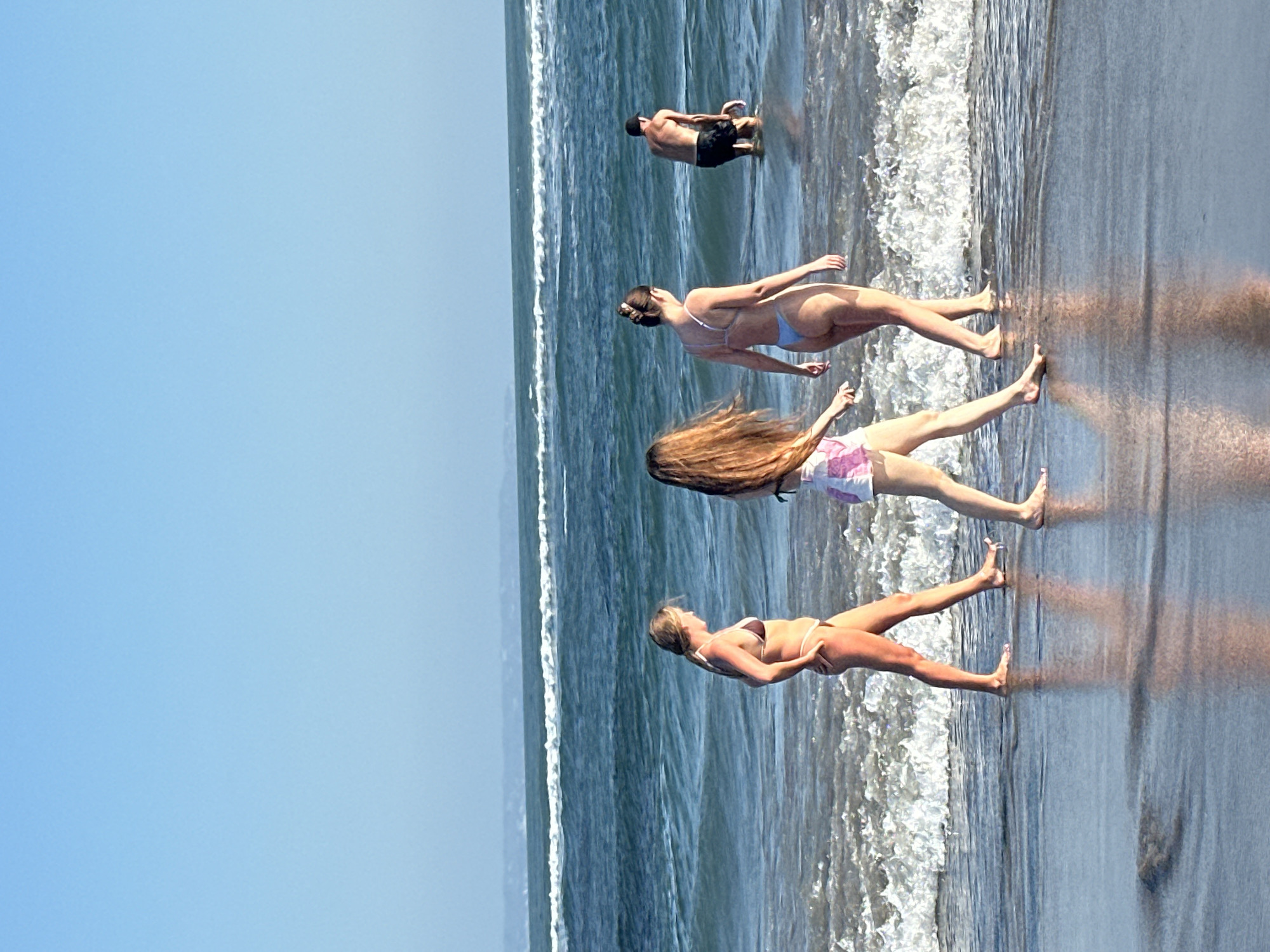 In the city of North Kuta, Indonesia, on a beach with calm, blue ocean waves and a clear blue sky, three women and one man are visible. Two of the women, dressed in bikinis, walk along the shoreline, their lower bodies submerged in shallow water. A third woman, also in a bikini, walks beside them. The man, wearing dark swim trunks, stands in the water further out, appearing to interact with a frisbee or similar object. The sand is wet, reflecting the sunlight, and gentle waves create a foamy white line along the beach.