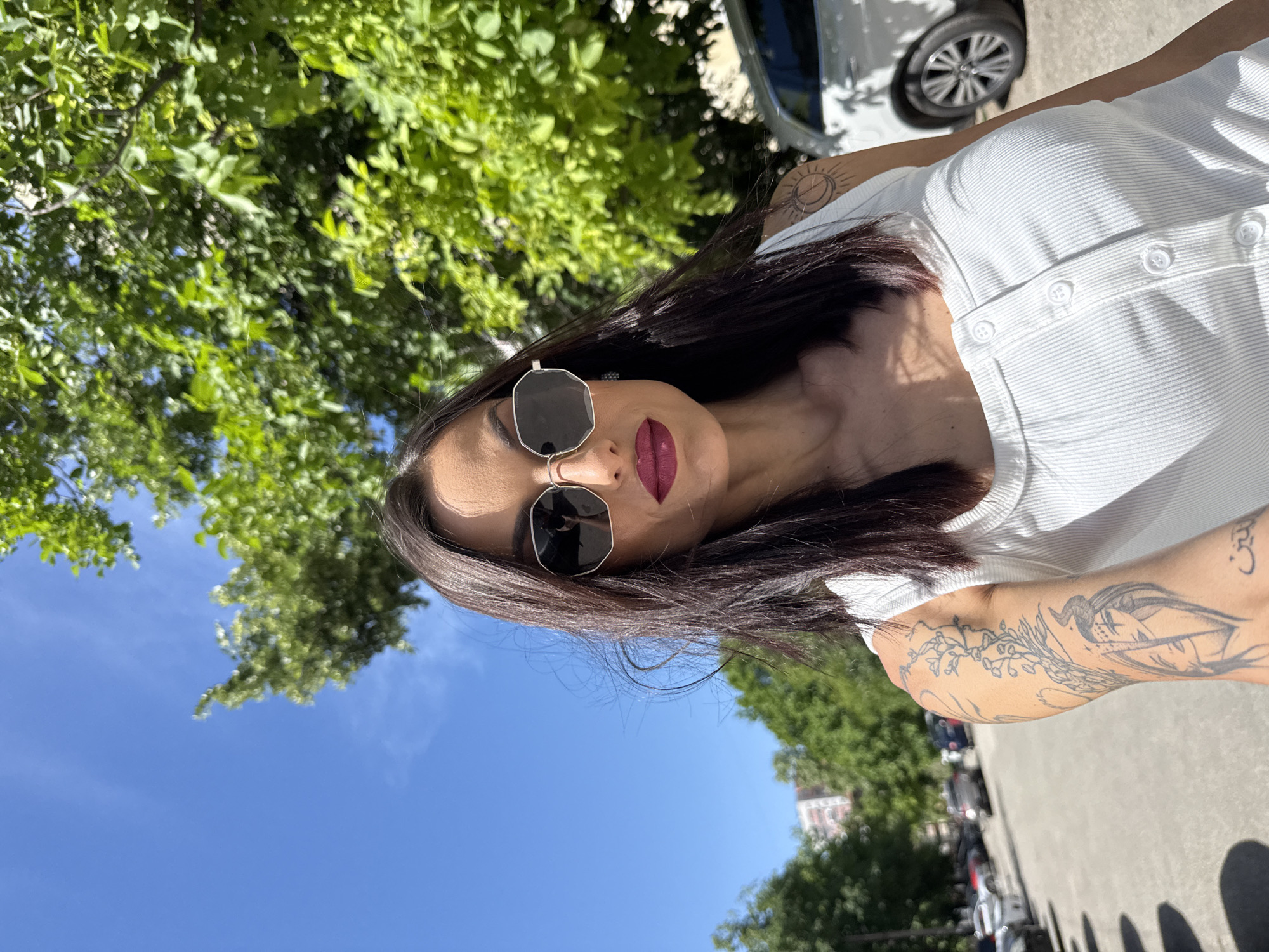 A medium-shot photo shows a woman with long, dark hair and burgundy lipstick. She wears black sunglasses and a white, buttoned top. Tattoos are visible on her arms. The background features green trees and a blue sky, suggesting an outdoor location. A portion of a white car is also visible. The location appears to be a city street lined with trees and parked cars. While the image is typical of an outdoor scene, there's no specific information to confirm the location as Pleven, Bulgaria.