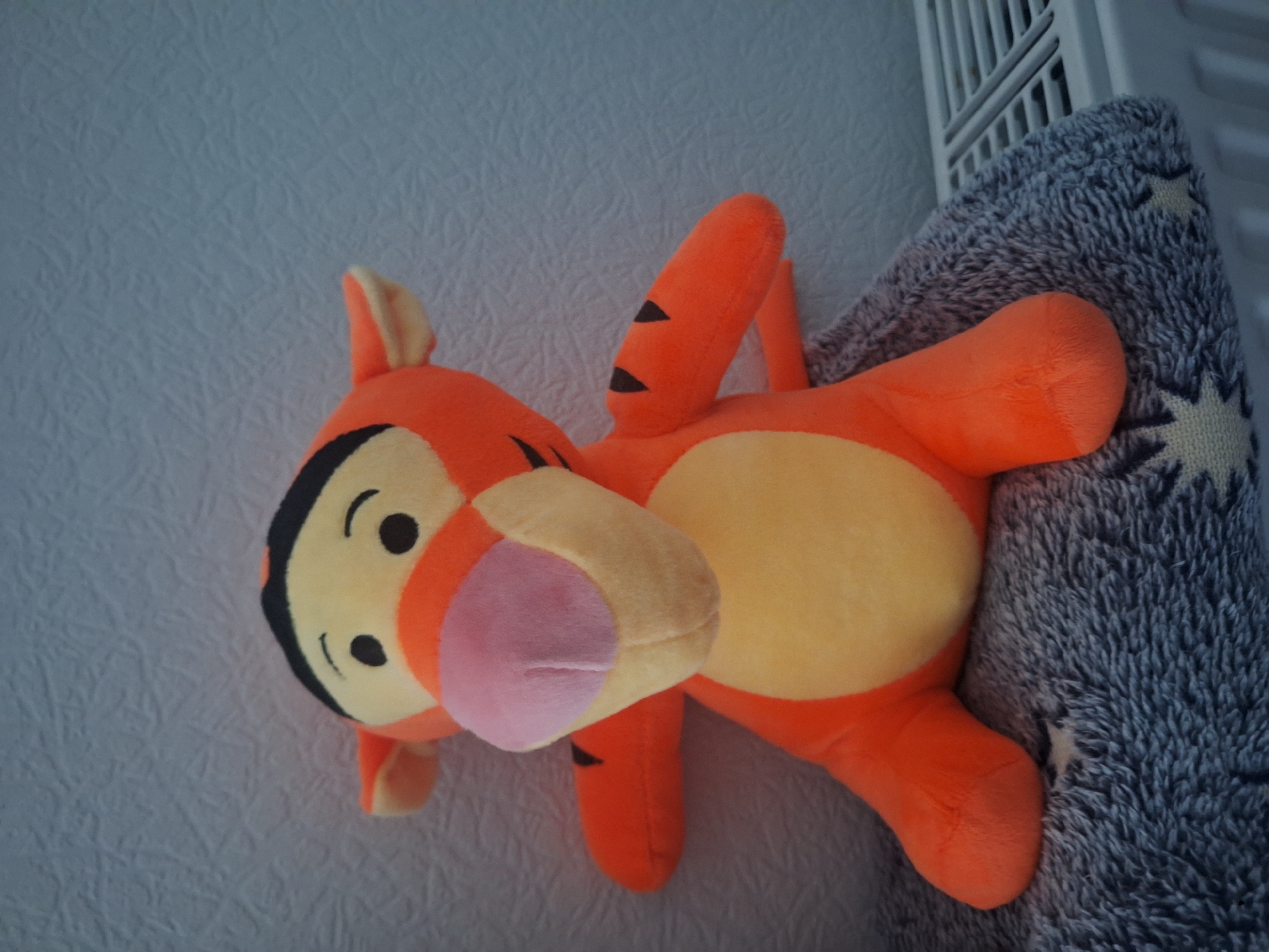 A plush toy Tigger, a character from Winnie the Pooh, is positioned on a grey, fuzzy blanket with a star pattern. The toy is orange with black stripes, a yellow belly, and a pink nose. It is sitting in front of a textured, light blue wall. To the right of the toy, a white radiator with horizontal bars is visible. The scene appears to be indoors. Pargolovo, Russia is a settlement, not a city.