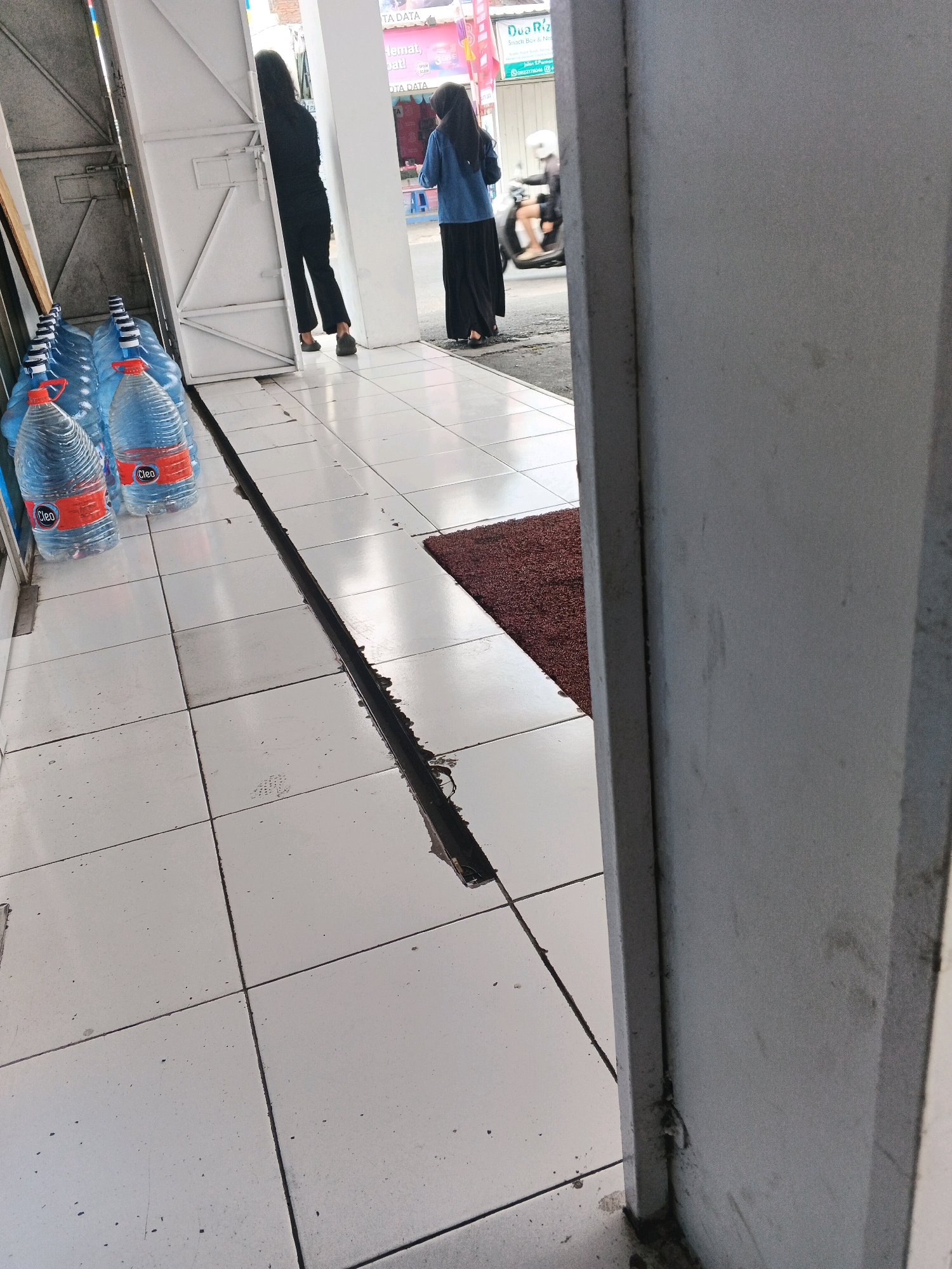 This is a street-level, eye-level shot of a sidewalk in Genuk Barat, Indonesia. The foreground features a row of large, clear plastic water bottles with red caps, branded "Cleo." Behind the water bottles, the sidewalk is tiled with white squares, divided by dark grout lines. A dark, narrow trench runs along the grout line between two rows of tiles. To the left, a white metal door is ajar, revealing the interior of a building. Two women are visible through the doorway. One, wearing black attire, stands facing away from the camera, partially obscured by the door. The other, wearing a blue top and a black hijab, stands further into the building, facing the camera. In the background, the street is visible, with a motorcycle rider wearing a helmet moving away from the camera. A sign with text is also visible on a building in the background. A maroon doormat is placed on the tiled sidewalk to the right of the water bottles. The overall lighting suggests daytime.