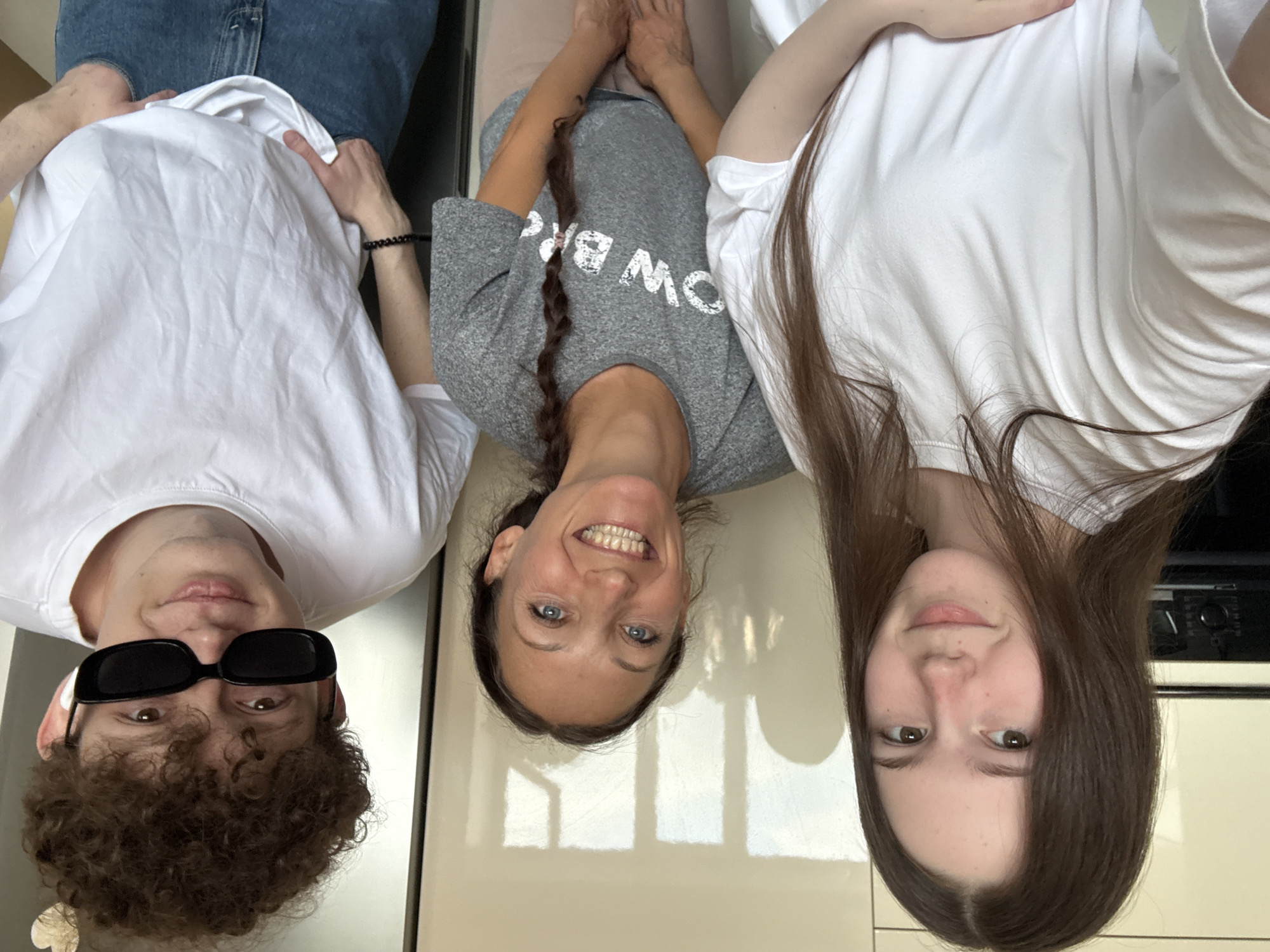 Three people are positioned from left to right, appearing to lie down and look directly into the camera. The person on the left wears a white T-shirt and dark sunglasses, and has brown curly hair. The person in the middle is wearing a grey T-shirt and has brown hair in a braid. The person on the right wears a white T-shirt and has brown hair. The background is a shiny, light-colored surface. The location and city are not discernible from the image.