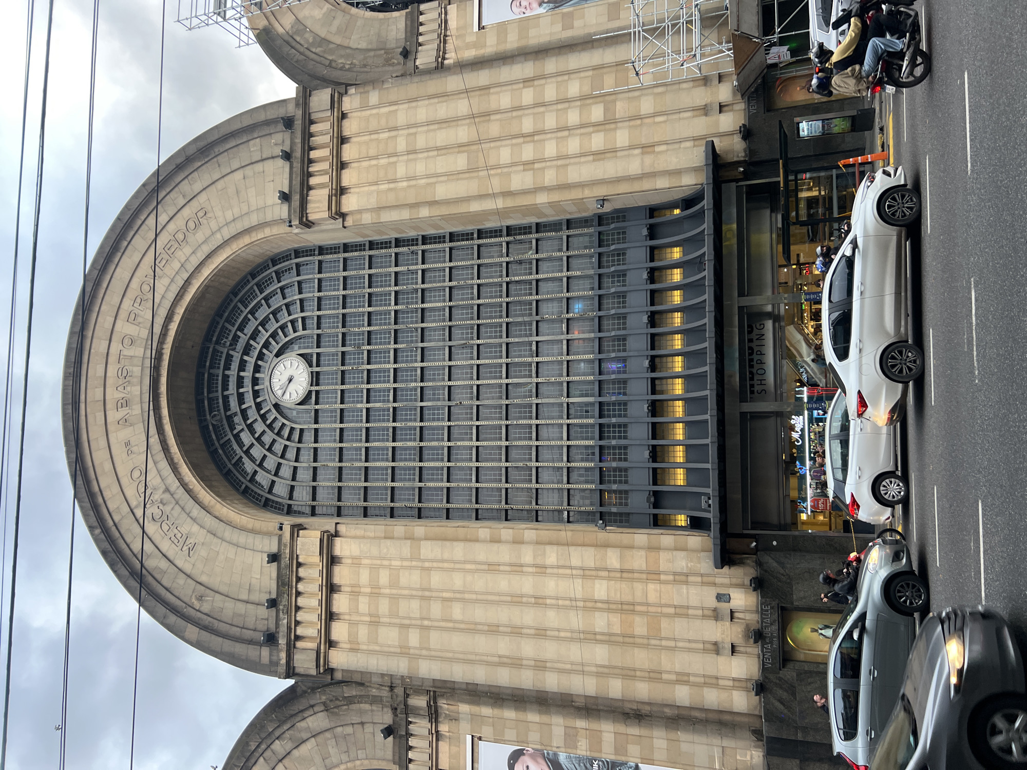 Here's a description of the image:
The image features the Abasto Shopping mall in Buenos Aires, Argentina, which was converted from a historic wholesale produce market. The facade is visible, characterized by its large arched window containing a clock. The building is made of light-colored stone or concrete.
Several vehicles are visible on the street in front of the building, including a motorcycle with two people on it, a white car, and a gray car. A group of people stands near the entrance, suggesting shoppers entering or exiting the mall. Signage for stores within the mall is visible above the entrance.