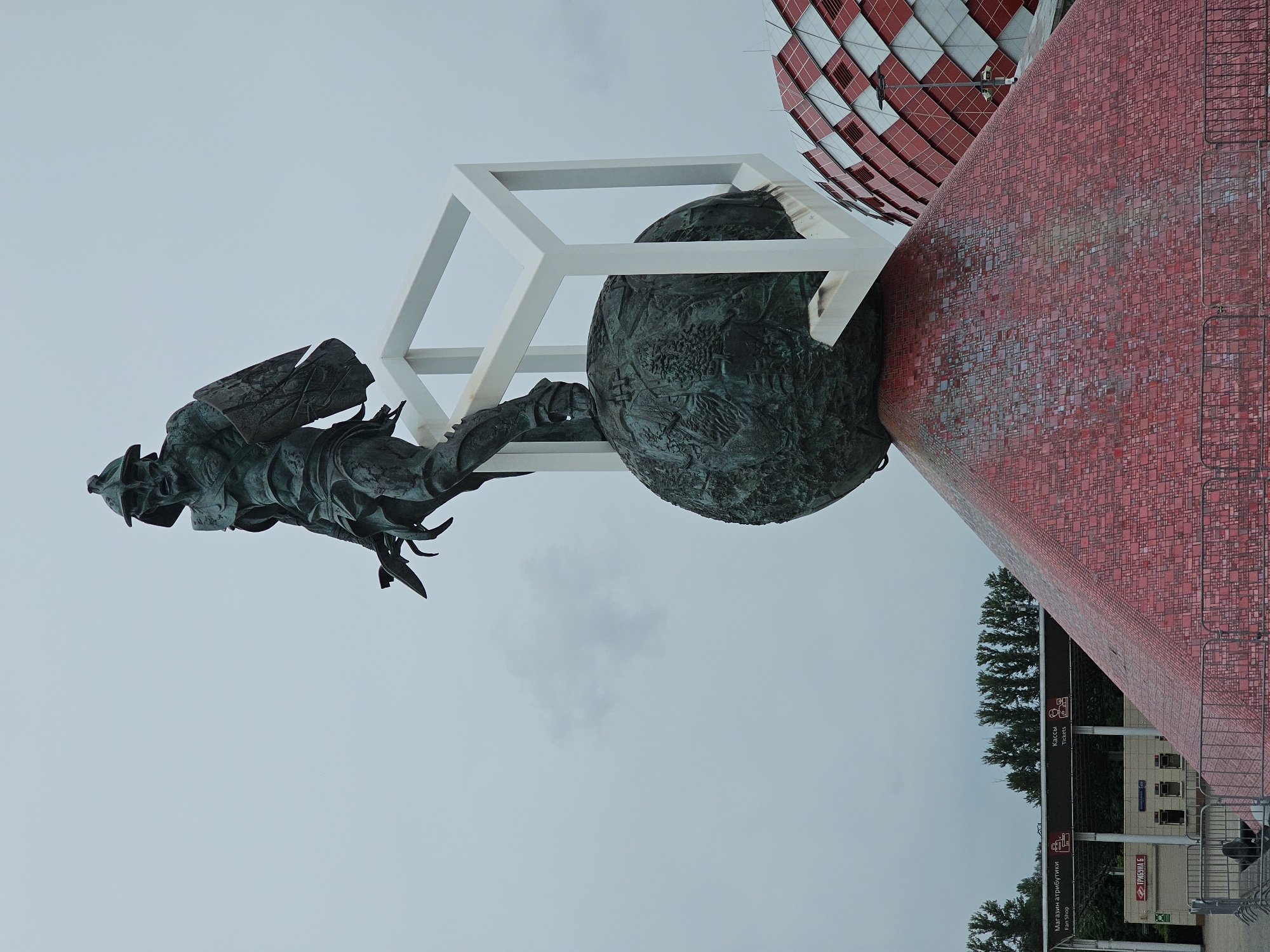 The image shows a statue of a gladiator atop a globe, which is itself placed on a red, pyramid-shaped pedestal. The statue is made of dark metal and depicts the gladiator in a dynamic pose, holding a shield and a sword. The globe is intricately decorated with relief carvings. A white, cubic frame structure surrounds part of the globe. In the background, there is a modern stadium with a distinctive red and white checkered facade. To the left of the pedestal, there are signs indicating a "Fan Shop" and "Tickets" in Russian, as well as a building with the inscription "ТРИБУНА 5" (Tribune 5). Trees are visible behind these structures. The sky is overcast. The overall scene is outdoors, likely in a public space or near a sports facility, in the city of Khimki, Russia, given the presence of the stadium and Russian signage.