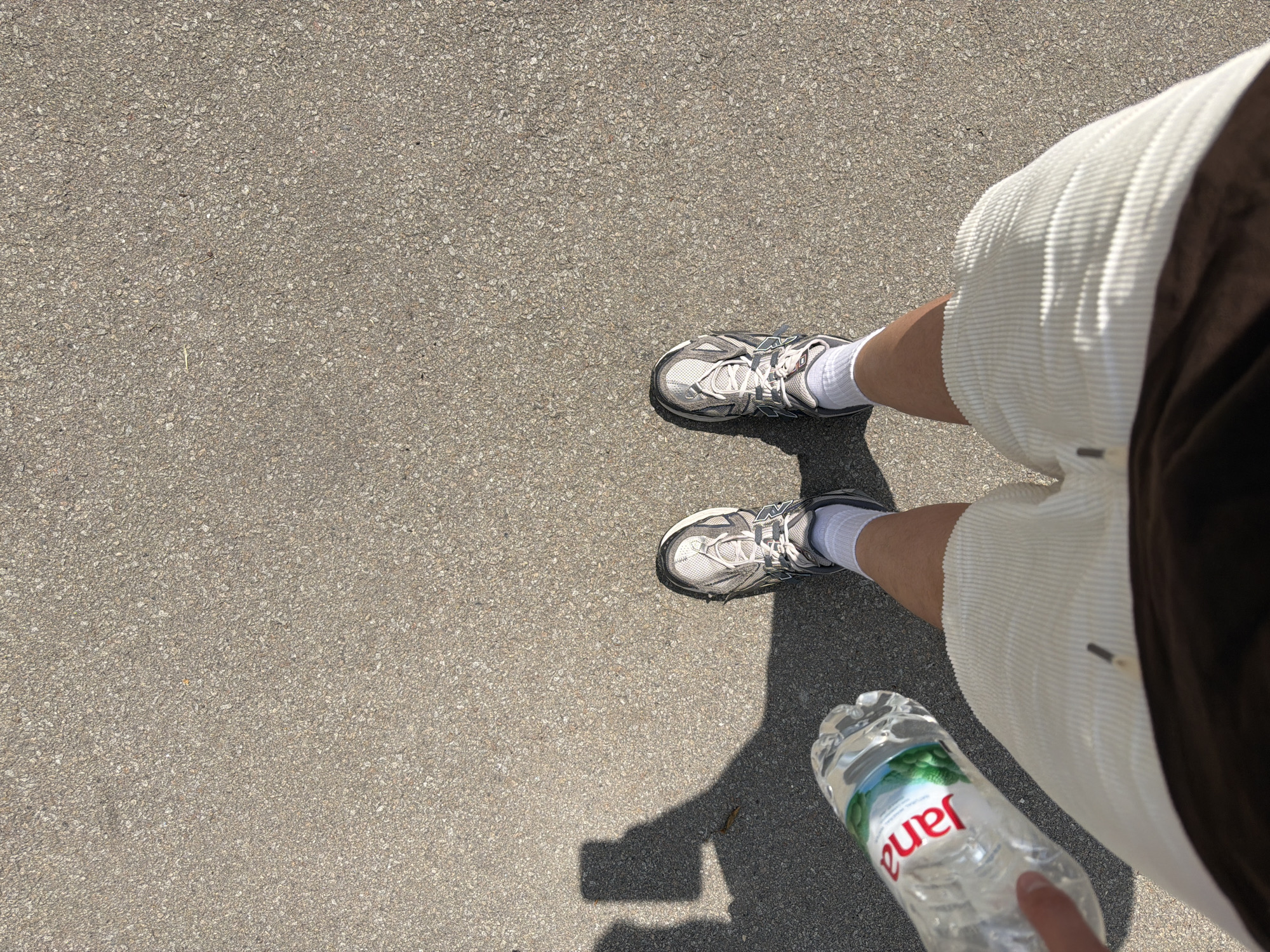 An eye-level, top-down shot captures a person standing on gray asphalt. The individual wears white athletic shoes with white socks and light beige corduroy shorts with a brown accent. The person's right hand holds a clear plastic water bottle labeled "Jana." The location is unknown, but no identifying signs point to the city of Belgrade, Serbia.