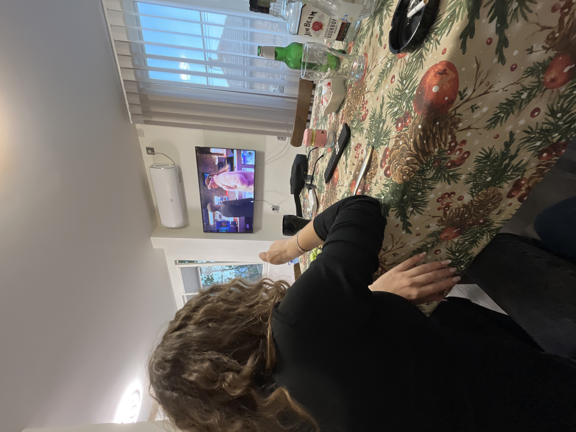 The image captures an indoor scene from a slightly elevated perspective, rotated clockwise. In the foreground, the back of a person, likely a woman with long, curly brown hair, is visible as she sits at a table. She wears a black long-sleeved top, and her left hand rests on the table.
The table is covered with a festive tablecloth featuring a pattern of pinecones, red berries, and green evergreen branches, suggesting a holiday or winter theme. On the table, various items are scattered: a bottle of "JIM BEAM" "KENTUCKY STRAIGHT BOURBON WHISKEY" (40% alc./vol.), an empty green glass bottle, a clear drinking glass, a small white dish, a hairbrush, reading glasses, a black remote control, a small pink container, and a black ashtray with a lit cigarette.
In the background, a flat-screen television is mounted on the wall, displaying a scene with two individuals, one in light attire and another in dark. To the left of the TV, a white wall-mounted air conditioning unit is visible. Above and to the right of the television, a window with horizontal blinds allows bright daylight into the room, suggesting a daytime setting. Another window further to the right offers a glimpse of outdoor greenery. The scene depicts a relaxed environment, possibly during leisure time, with the person enjoying drinks and watching TV.