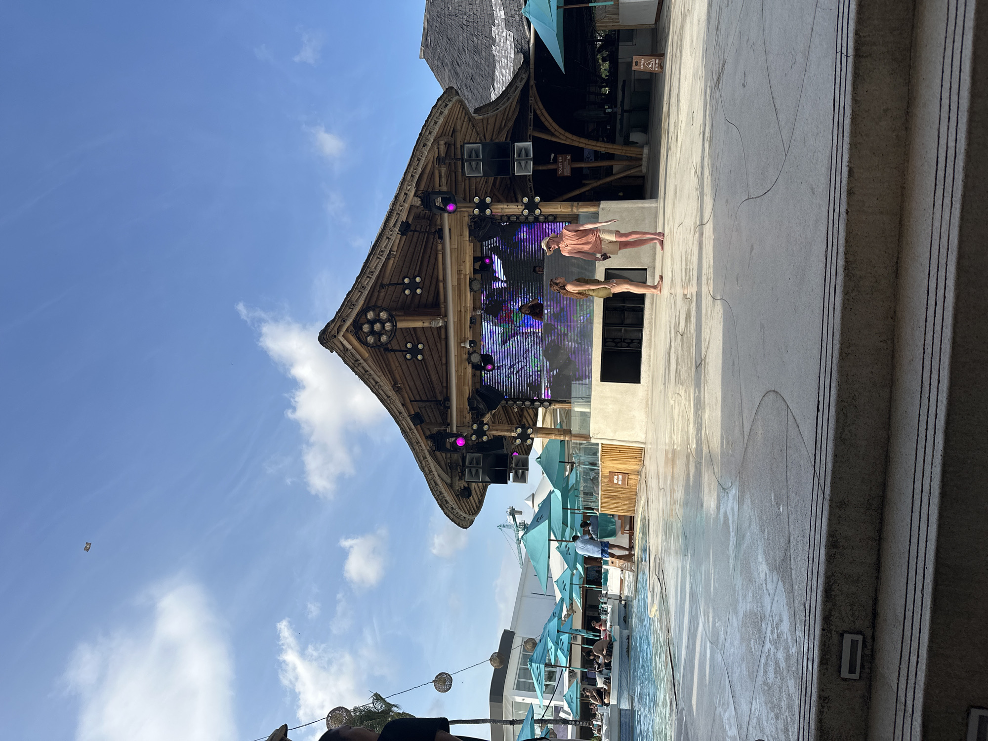 The image captures a scene at a venue, likely a beach club, in North Kuta, Indonesia. The architecture features bamboo structures, including a large stage with sound equipment and lighting. A large screen displays vibrant graphics. Two individuals, a man and a woman, are standing on the stage. Numerous blue umbrellas line a pool area, with people visible in the background, indicating a leisure environment. The sky is blue with scattered clouds. The foreground shows stone flooring or steps leading to the venue.