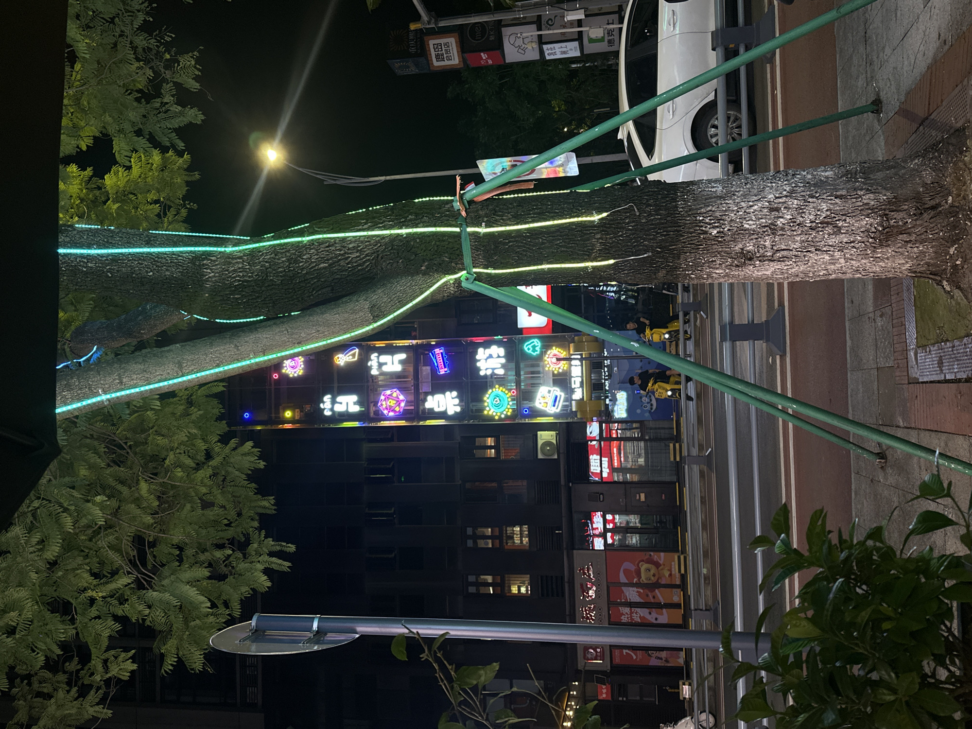 In the city of Chongqing High-tech Zone, China, at night, a tree is illuminated with green and blue LED lights wrapped around its trunk and supported by green metal poles. The tree is on a sidewalk adjacent to a street with traffic lights and road markings. In the background, several buildings with illuminated signs and windows are visible. One building has a tall vertical sign with various neon lights, including Chinese characters and colorful shapes. Across the street, businesses with illuminated storefronts are present. A white car is parked on the right side of the road, and a person on a yellow scooter is riding on the left side of the street. Green foliage is in the foreground. The scene captures a nighttime urban environment with commercial activity and decorative lighting.