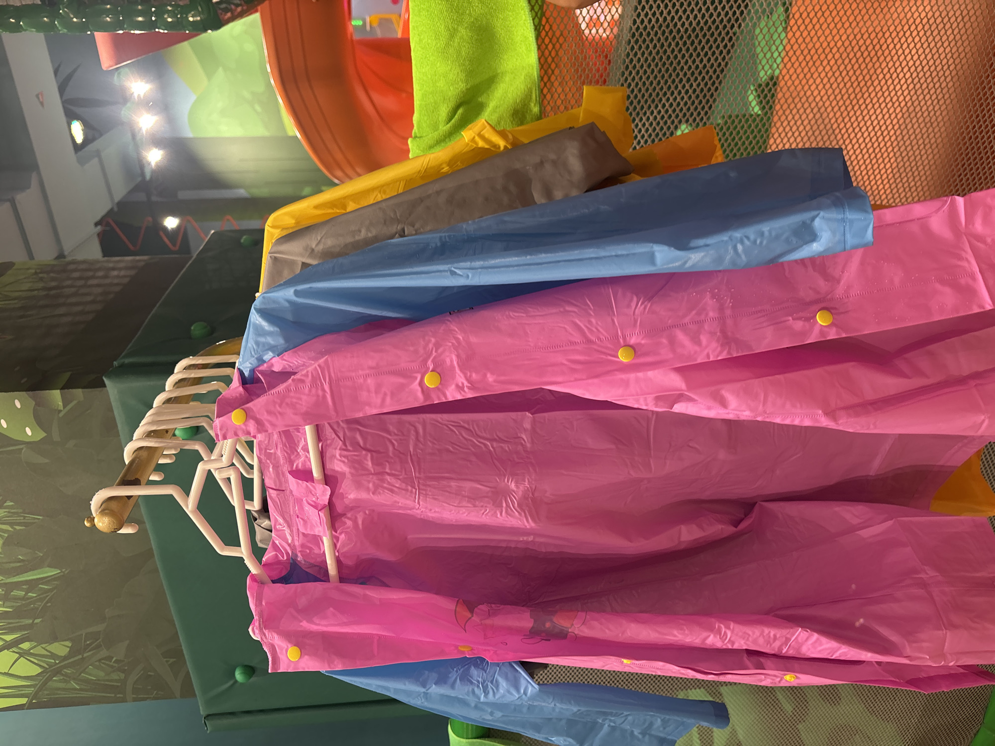 The image shows a close-up of several colorful raincoats hanging on a rack. The raincoats are pink, blue, gray, and yellow. They appear to be made of a plastic material and some have yellow buttons. The background suggests an indoor play area or amusement park with vibrant green and orange structures, possibly a playground slide. The lighting is bright, with some spotlights visible in the upper background. The scene is clean and orderly. There are no people visible in the image. The overall impression is that of a child-friendly environment. The location in Dubai, United Arab Emirates is not specified by any visual cues in the image itself.
