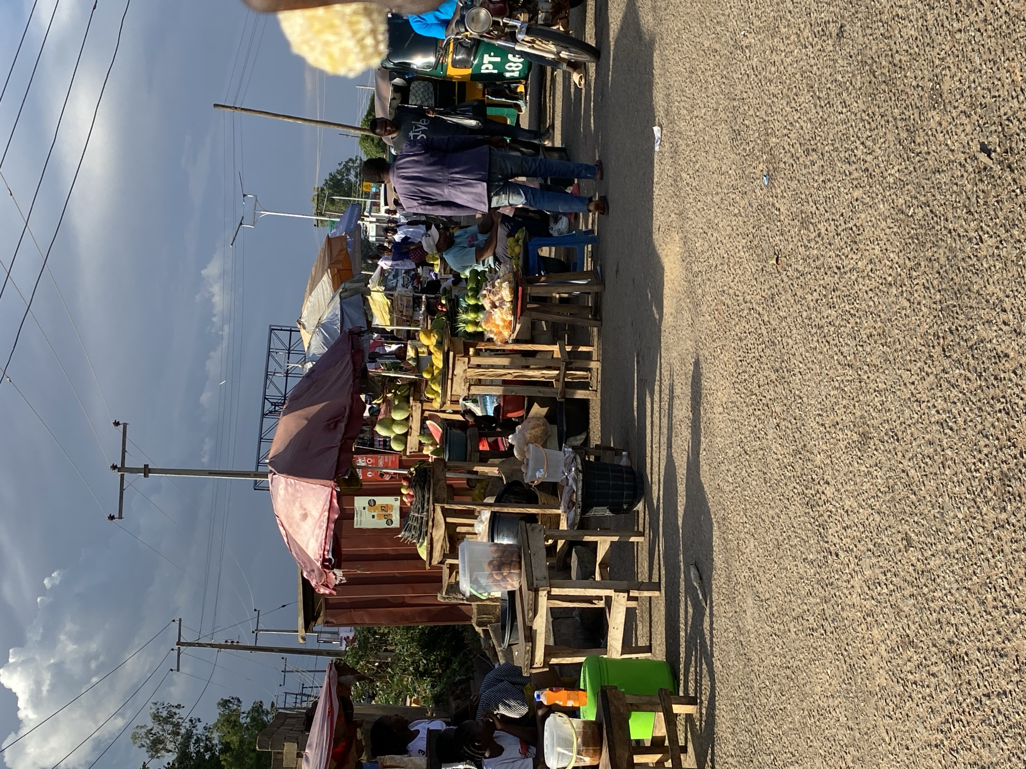 The image depicts a street scene in Jos, Nigeria, lined with makeshift stalls selling various goods. People are gathered around the stalls, appearing to be shopping or conversing. Fruits like pineapples and coconuts are visible on display. The stalls are constructed from wood and covered with tarps or corrugated metal sheets. A "keke napep" (auto rickshaw) is parked on the side of the road, and power lines run above the stalls. The road is paved, and shadows indicate the time of day is likely mid-morning or mid-afternoon.