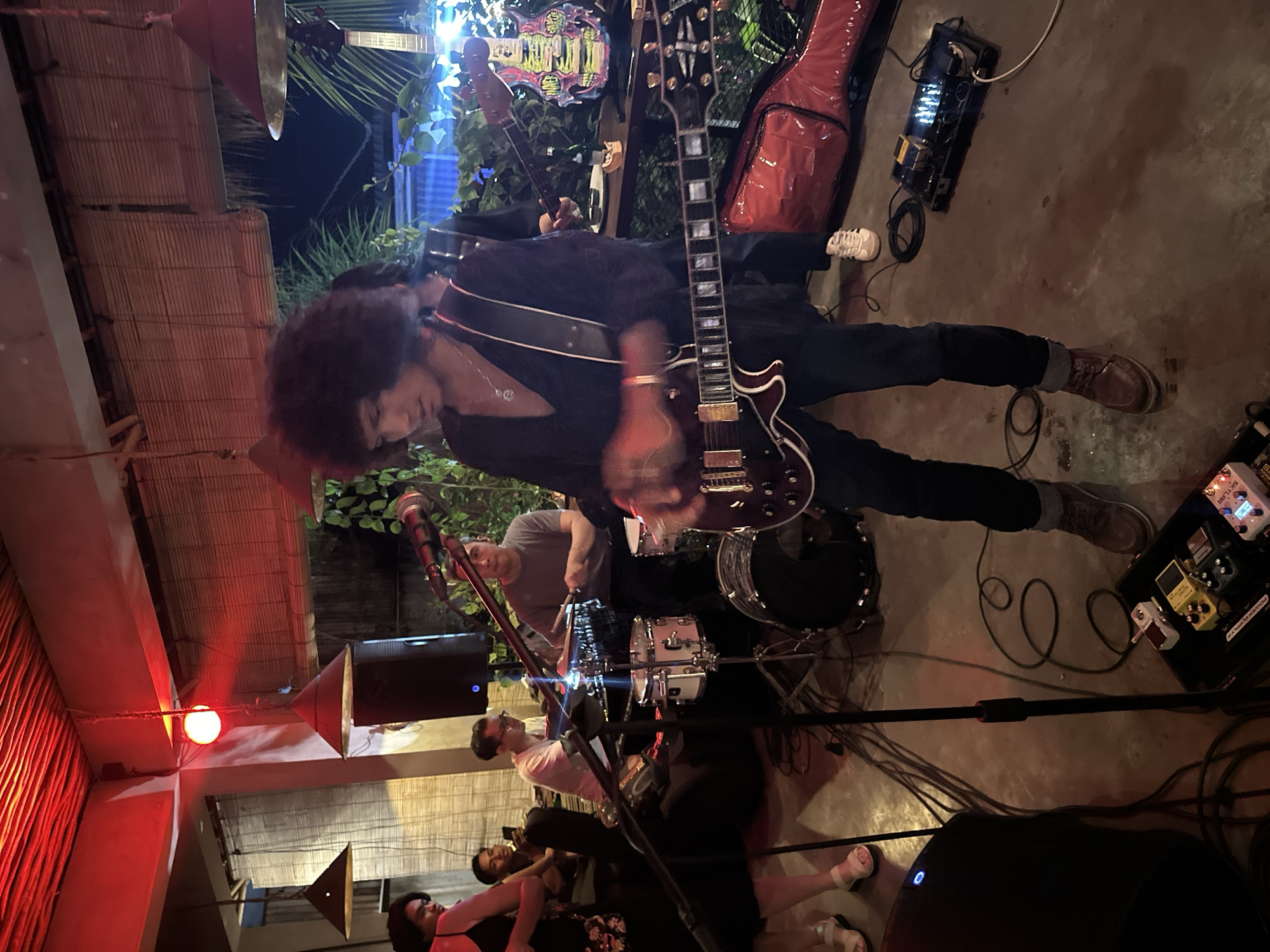 The image depicts an indoor performance space, possibly a bar or restaurant, in Kuta Selatan, Indonesia. Red lighting illuminates the ceiling, while bamboo blinds cover windows or walls. A band is performing, with a lead guitarist in the foreground playing a dark-colored electric guitar and wearing a black open-neck shirt and dark pants. He has a large, curly hairstyle and a necklace. To his left, a drummer is seated behind a drum kit, wearing a gray t-shirt. Other band members and audience members are visible in the background. Audio equipment, including microphones, speakers, and a pedalboard with multiple effect pedals, are arranged on the floor. A red guitar case is visible on the right side of the frame. The overall atmosphere suggests a live music event in a casual setting.