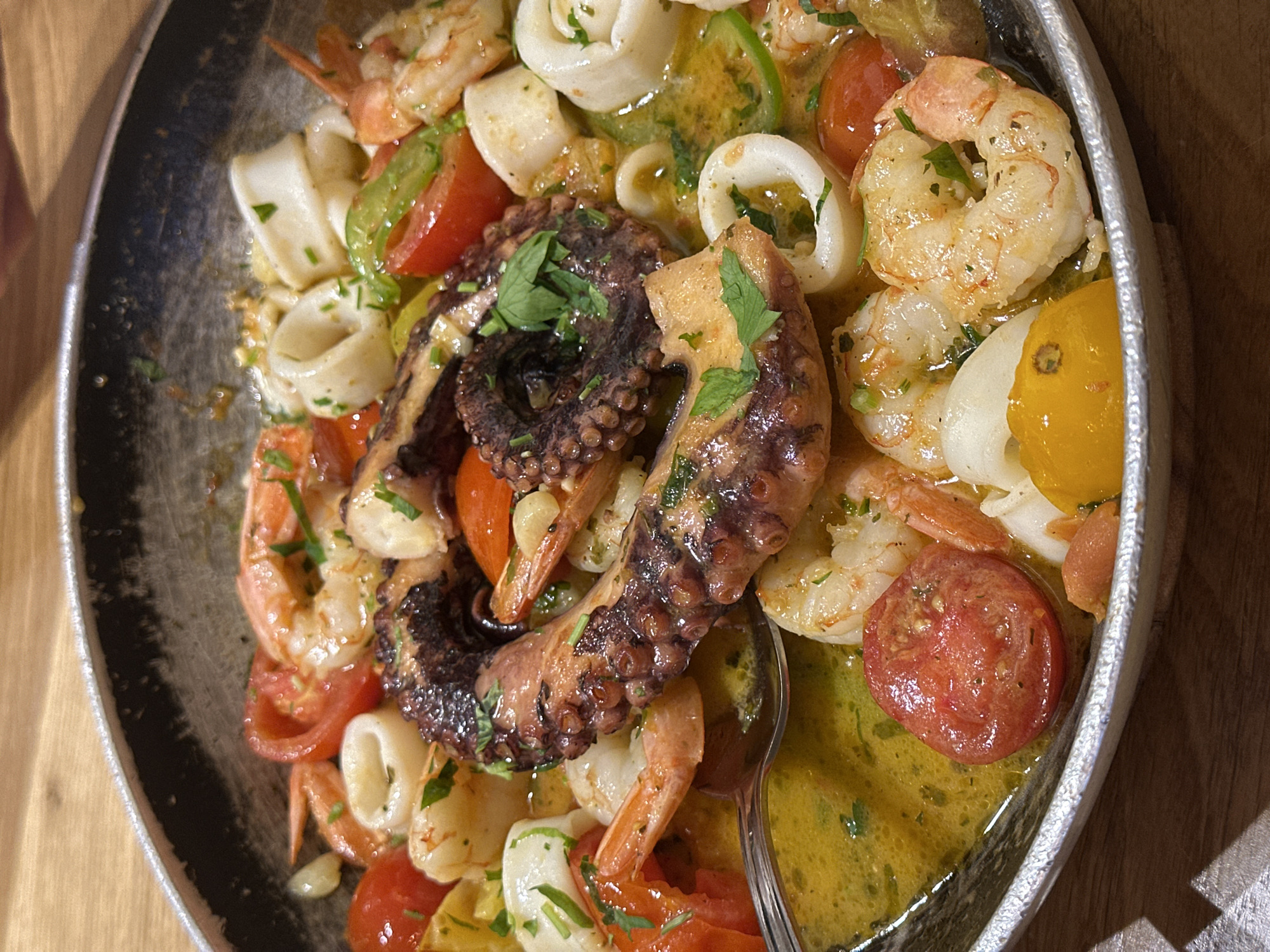 The image displays a close-up, overhead view of a seafood dish served in a rustic, shallow pan. The dish prominently features a large piece of cooked octopus tentacle as the centerpiece, surrounded by shrimp, calamari rings, cherry tomatoes (both red and yellow), and green peppers. The seafood is swimming in a vibrant, golden-yellow broth, glistening with oil and flecked with chopped parsley. A metal spoon is visible in the lower left portion of the frame, partially submerged in the broth, suggesting the dish is ready to be eaten. The pan itself appears to be made of metal with a slightly textured, dark interior and a rim that shows some wear. The dish is placed on a wooden surface, likely a table, with a visible wood grain. The lighting suggests an indoor setting, possibly a restaurant. The specific location of Tel-Aviv, Israel is not indicated by any visual cues within the image itself.