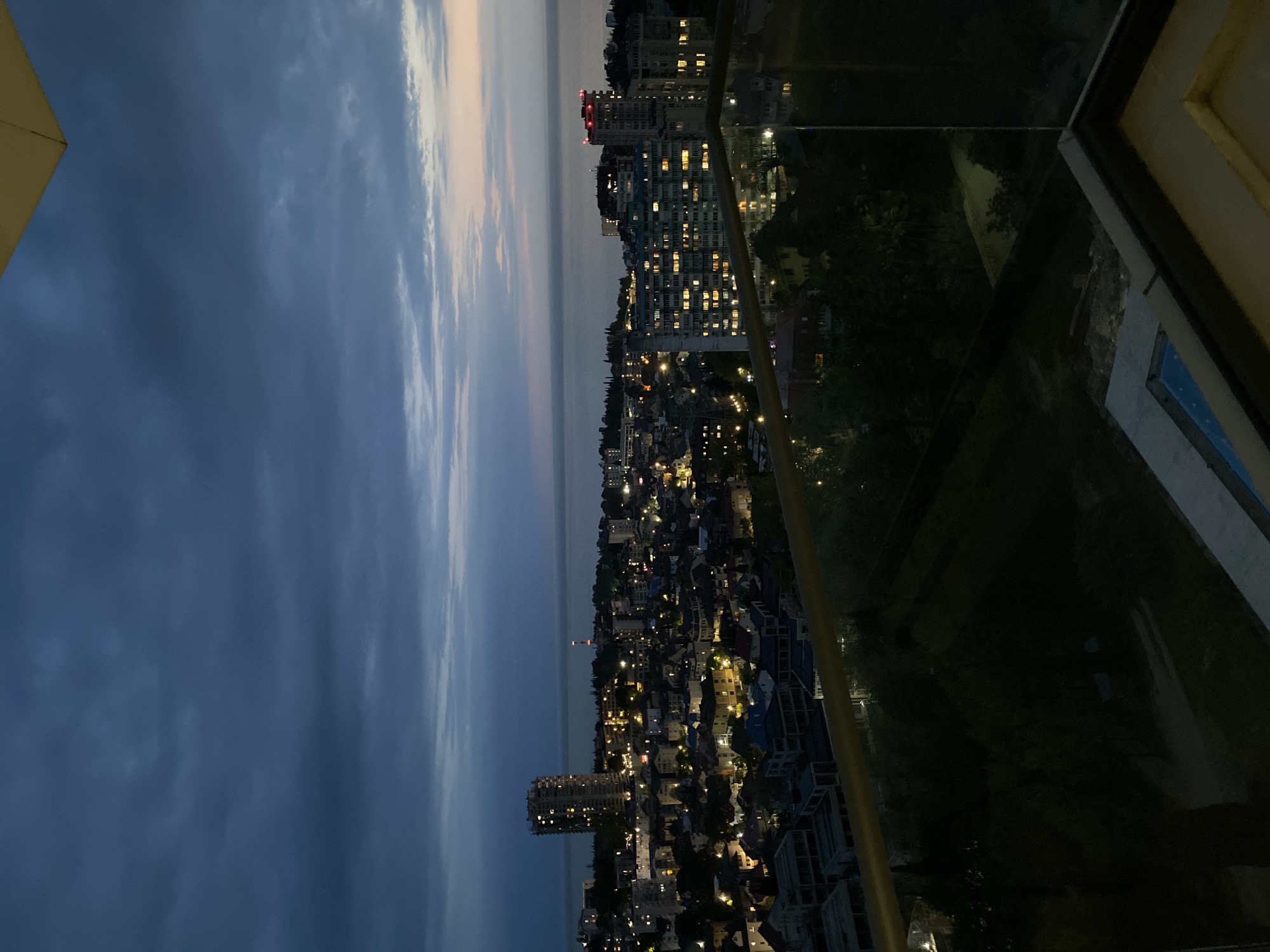 A dusk-lit cityscape, possibly Sochi, Russia, is viewed from an elevated perspective, likely a balcony or high-rise building. The sky transitions from dark blue to pale orange above the horizon, indicative of sunset or sunrise. Coastal water is faintly visible at the horizon. A dense urban environment unfolds below, comprising numerous buildings with illuminated windows, suggesting residential or commercial occupancy. Clusters of lights form a network of roads and structures. The foreground includes parts of the balcony from which the photo is taken. A glass panel and railings are partially visible, indicating the boundary of the viewing point. The atmosphere appears calm and still, marked by the transition between day and night.