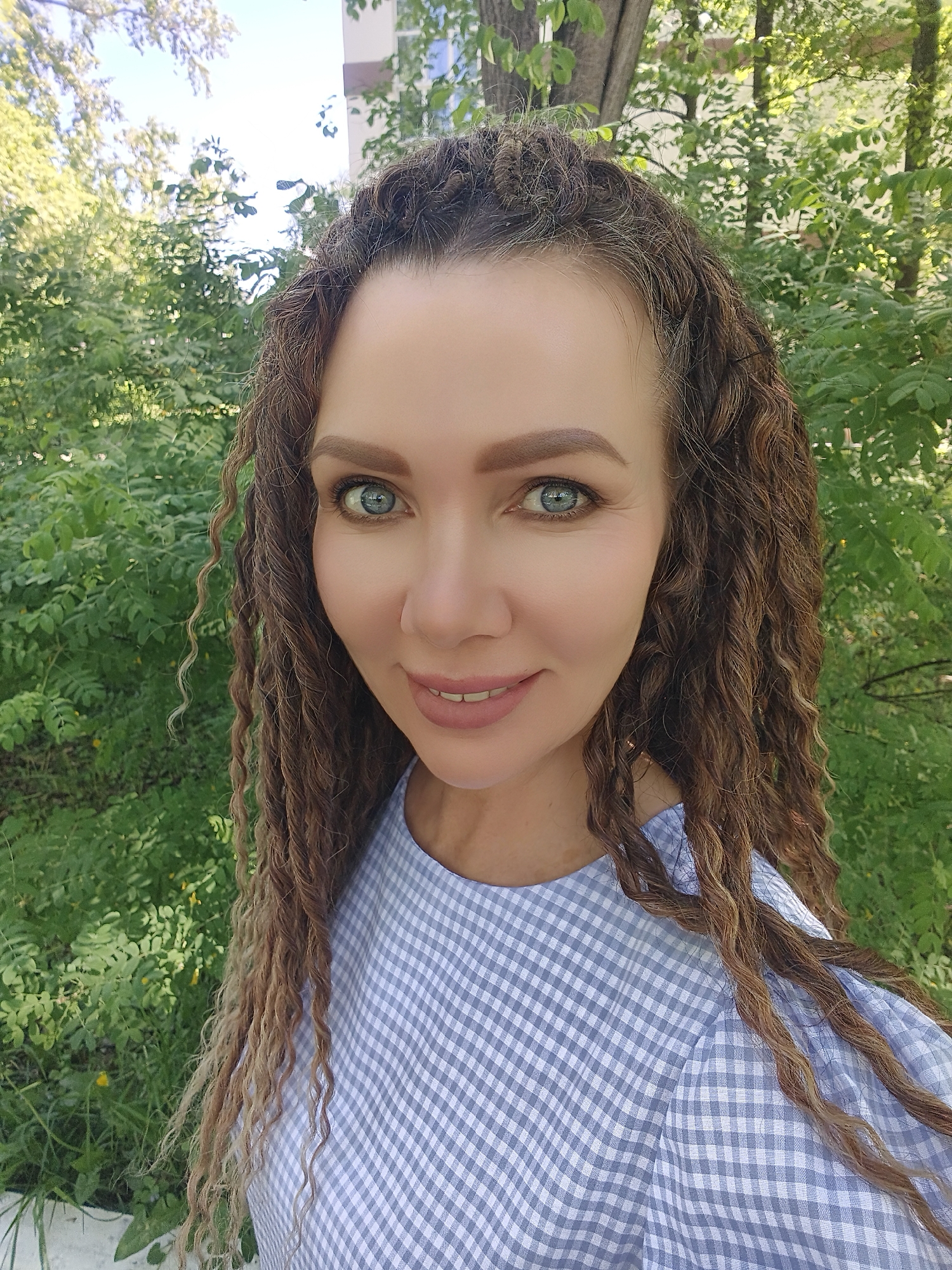 A woman with long, curly brown hair is smiling at the camera. She has blue eyes and is wearing a blue and white checkered shirt. The background is a lush green garden or park.