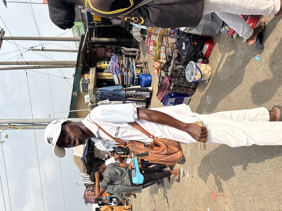 A man wearing a white shirt, white pants, a white baseball cap, and a brown leather shoulder bag stands in a crowded outdoor market. He holds what appears to be a stack of money in his left hand. In the background, vendors display various goods, including clothes, fabrics, and packaged food. Wires hang overhead, and other people are walking through the market.