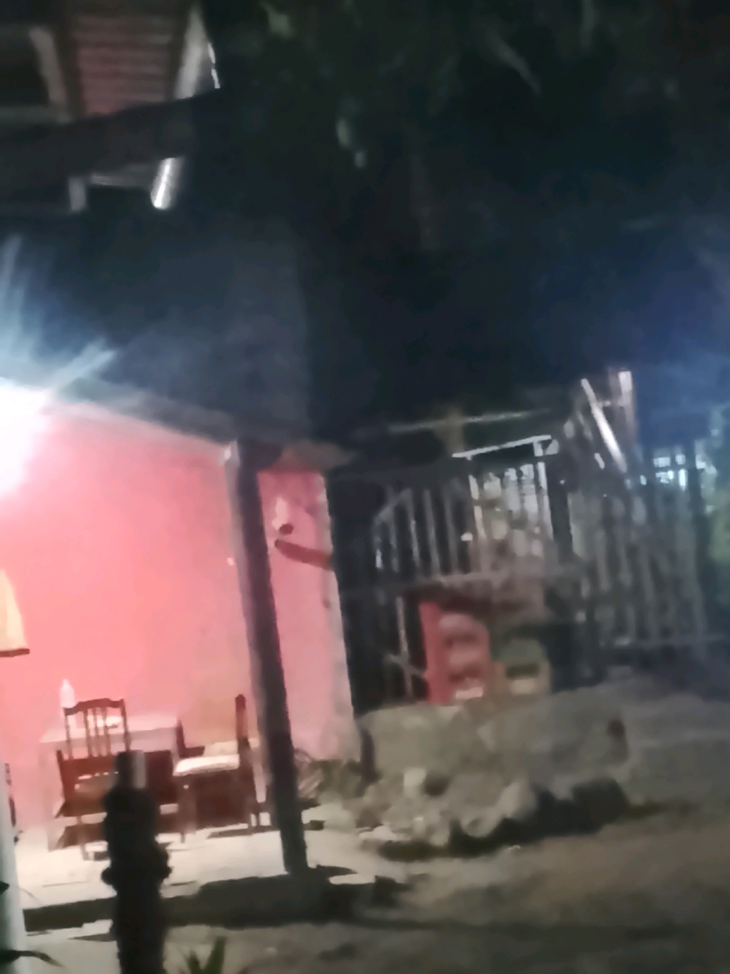 A nighttime outdoor scene in Ujungrusi, Indonesia, shows a red-walled building with a covered porch. Two wooden chairs and a table are visible under the porch. To the right, a structure with vertical bars and a metal roof appears to be some kind of enclosure or fencing. The ground is dirt and debris. Lighting is dim, with some artificial lights creating bright spots and casting shadows. The image is blurry, suggesting it was taken in low light conditions or with a moving camera.