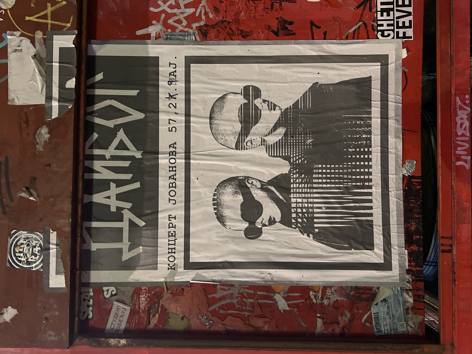 The image shows a poster adhered to a weathered, red metal surface, possibly a bulletin board or a door, in an urban outdoor environment. The poster itself is largely monochrome, featuring two stylized, bald figures wearing large, dark sunglasses, one slightly behind the other. Their faces are rendered in a graphic, almost pixelated style. Above the figures, in bold, stylized Cyrillic lettering, appears the word "ДАНБОГ". Below the figures, in smaller Cyrillic text, it reads "КОНЦЕРТ ЈОВАНОВА 57,2К. МАЈ". The poster is creased and slightly torn in places, suggesting it has been exposed to the elements.
Surrounding the central poster, other elements are visible. To the left, there are fragments of other posters and stickers, some showing peeling red paint underneath. One visible sticker has the word "ARTMORIA" and a QR code. To the upper left of the main poster, a circular sticker with an eye-like graphic is present. To the right of the main poster, red paint is heavily graffitied with black spray-painted lines and lettering. Two small stickers are visible on this red surface below the main poster, one with "GHETTI" and another with "FEVE". At the very bottom of the frame, a red metal bar is visible, with some graffiti in white marker.
The overall impression is that of urban street art and advertising, common in public spaces in Belgrade, Serbia, where Cyrillic script is used. The style of the poster and the surrounding graffiti suggest a specific artistic or cultural context.
