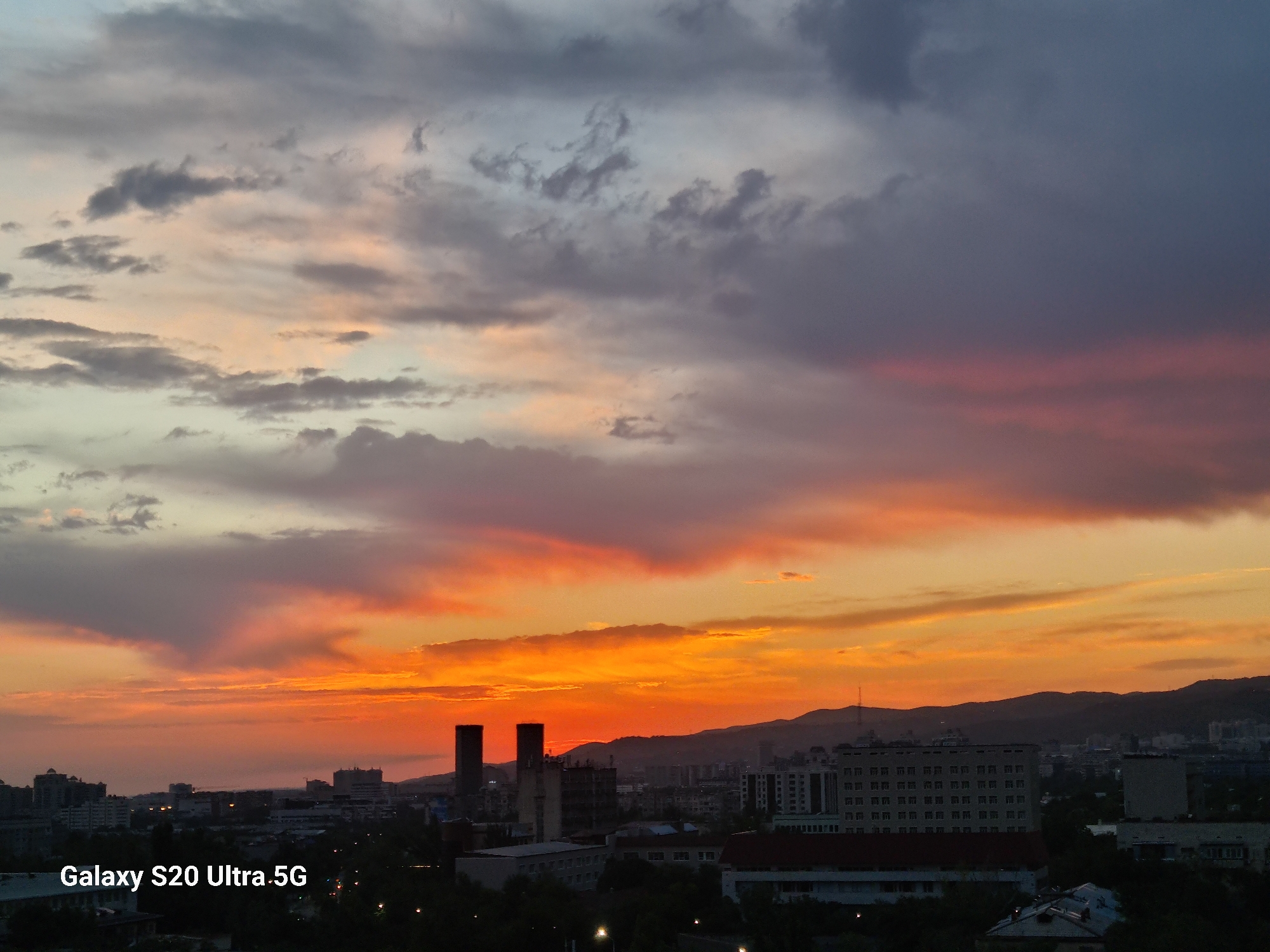 The image captures a vibrant sunset over the city of Almaty, Kazakhstan. The sky is ablaze with hues of orange, pink, and purple, with dramatic clouds adding texture and depth. Silhouetted against the colorful sky are the distinctive twin towers of the Kazakhstan Hotel, a landmark in Almaty. In the foreground, the cityscape is visible, with various buildings and hints of greenery. The lower left corner of the image contains a watermark: "Galaxy S20 Ultra.5G". The overall impression is a dramatic and beautiful urban landscape at dusk.