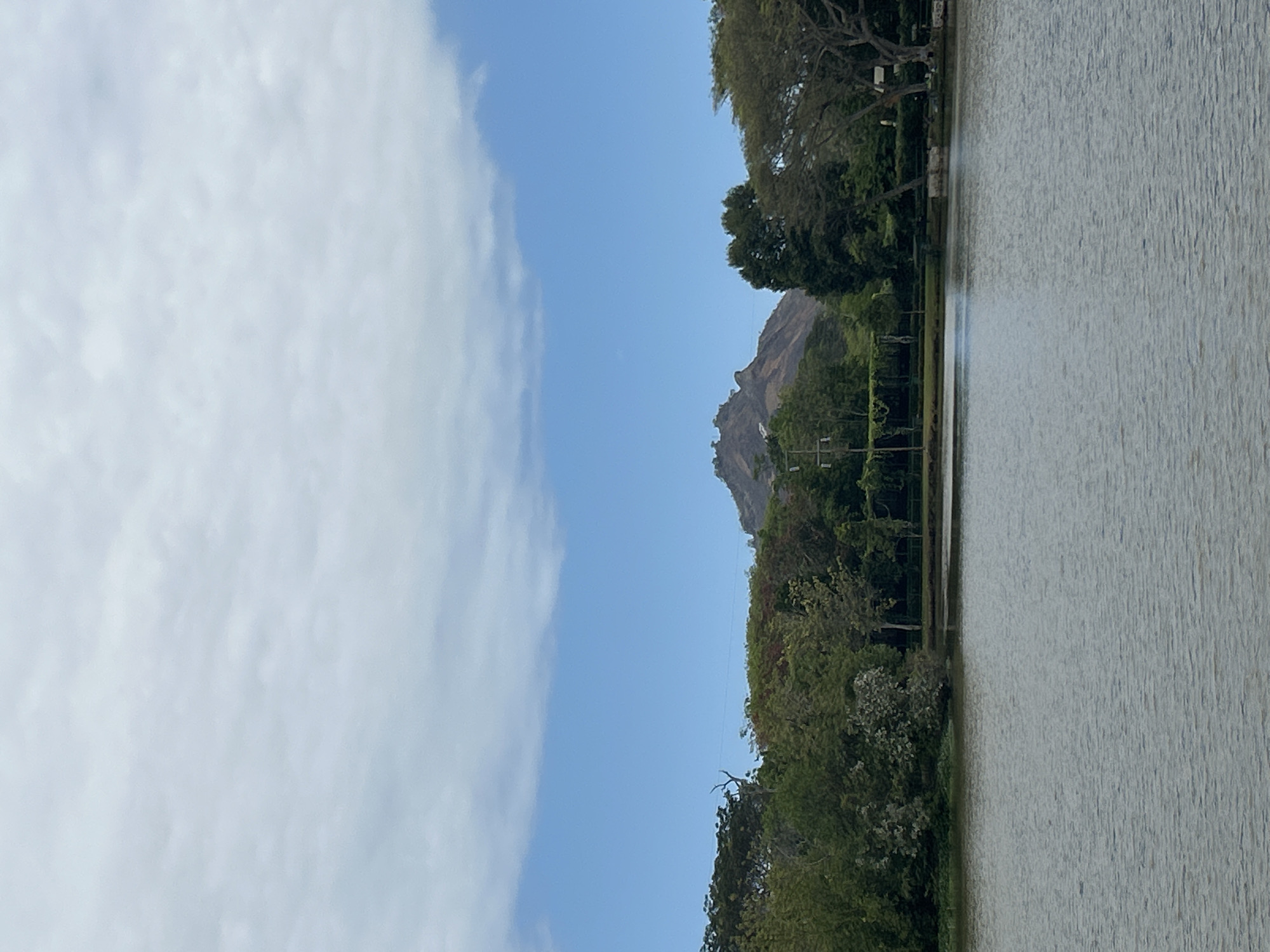 The image depicts a serene landscape with a body of water in the foreground. The water reflects the light, showing ripples on its surface. Behind the water, a lush green treeline runs across the middle ground, with various types of trees, some taller and denser, others more sparse. Interspersed within the trees are glimpses of what appears to be manicured lawns and some structures, possibly park benches or small gazebos, hinting at a maintained public space.
Dominating the background is a prominent, rocky mountain peak, characterized by its rugged texture and uneven surface. The mountain has a distinctive shape with several craggy outcrops and appears to be covered in sparse vegetation at its base.
The sky above is a vibrant blue, with scattered, soft white clouds that add texture and depth to the upper portion of the image. The lighting suggests it is daytime, likely mid-morning or afternoon, with diffused sunlight. The overall scene evokes a sense of tranquility and natural beauty, a common characteristic of outdoor recreational areas. The location is identified as Tanubada, Papua New Guinea.