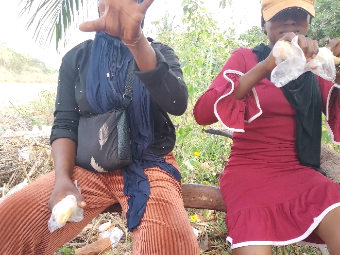 Two individuals are depicted outdoors in Ejule, Nigeria. The subject on the left, identified as female, is seated, wearing a black long-sleeved top with white embellishments, brown pleated pants, and a dark blue headscarf. A black shoulder bag with a white leaf pattern is slung across her body. Her right hand is raised with fingers splayed, while her left hand holds a small, light-colored object wrapped in clear plastic. The subject on the right, also identified as female, is seated next to her, wearing a red fitted dress with ruffled sleeves and hem trimmed in white, and a yellow baseball cap. She holds two similar light-colored, plastic-wrapped objects, appearing to unwrap one with both hands. Both individuals are positioned on a horizontal wooden log or branch. The immediate ground level consists of dry plant debris and some discarded white plastic. The background features sparse green vegetation and a partially visible dirt bank on the left, with a palm frond in the upper left.