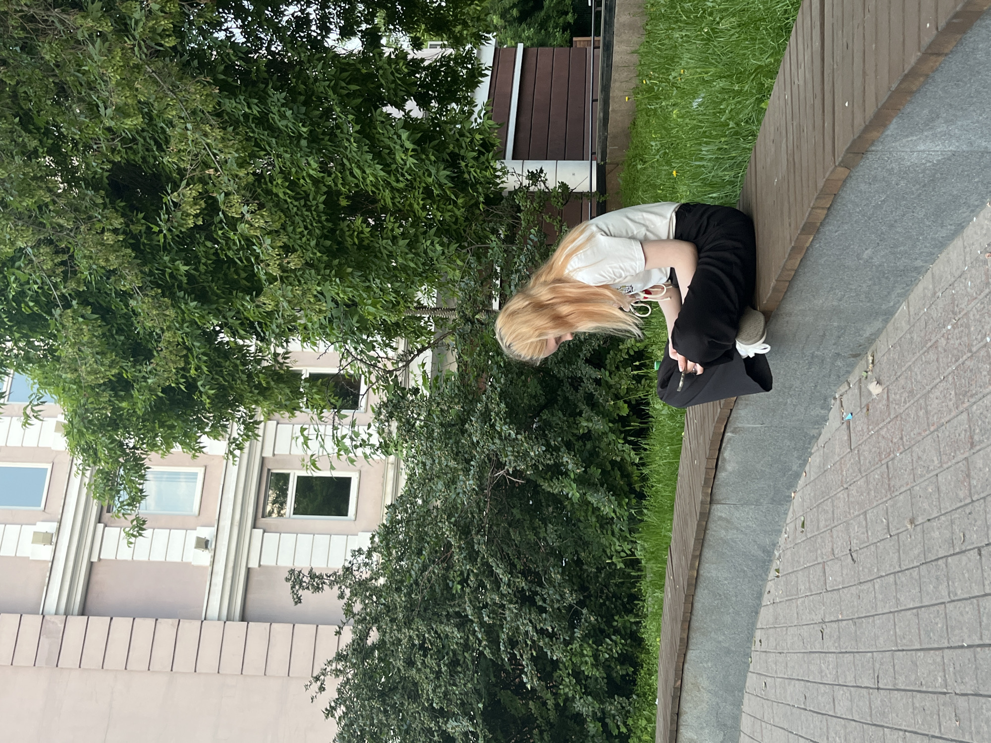 A young woman with blonde hair sits cross-legged on a curved wooden bench in a park setting, looking down at her phone. She is wearing a white t-shirt and black pants. The bench is situated in front of a grey stone retaining wall, with a paved area in the foreground. Behind the bench, a lush green lawn is visible, with a large tree providing shade. In the background, a pale pink building with white trim and windows stands adjacent to a dark brown wooden structure. The scene appears to be a public outdoor space in the city of Krasnoyarsk, Russia, possibly a park or a courtyard.