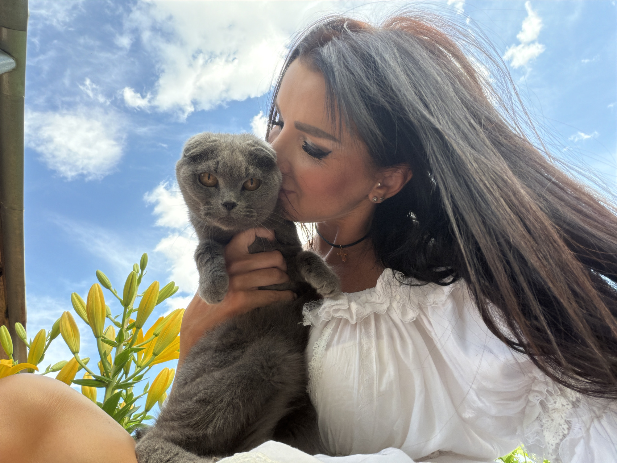 In Novaya Usman', Russia, a woman with long, dark hair and heavy eye makeup is captured from the chest up, embracing and kissing a gray Scottish Fold cat. She is wearing a white, ruffled blouse with lace trim. The cat is being held by the woman's arms. In the background, a bright blue sky with scattered white clouds is visible. To the left of the frame, a stalk of yellow lilies with unopened buds is present, and to the far left, a portion of a metal awning or roof edge can be seen. The overall scene suggests an outdoor setting, possibly a garden or patio, under a clear sky.