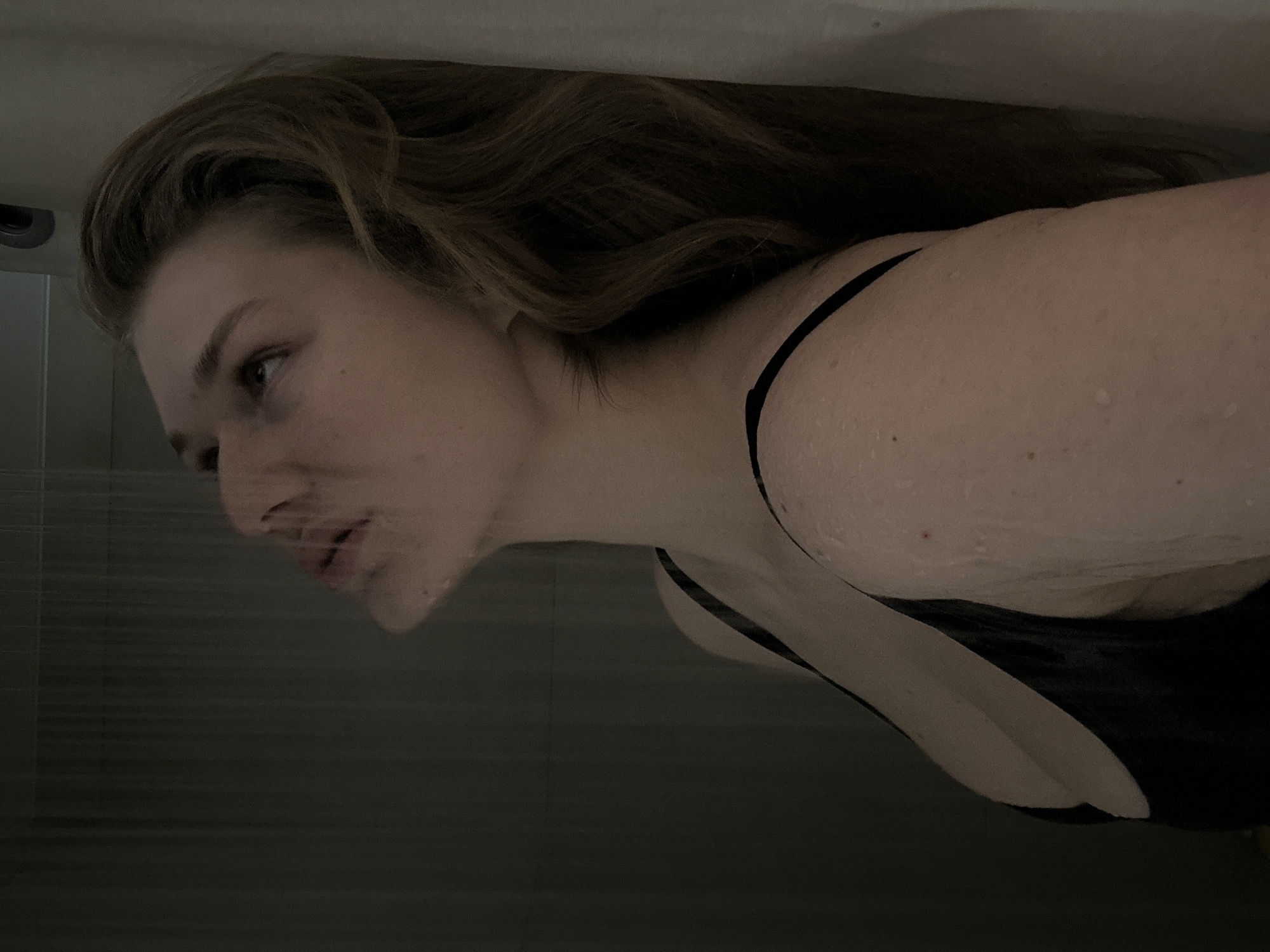 A woman is shown in a profile-view photograph. She appears to be in a shower, as evidenced by the wet skin and the tiled wall in the background. She has long, light brown hair. She is wearing a black top with thin straps. It is impossible to verify that the location is Kazan, Russia.