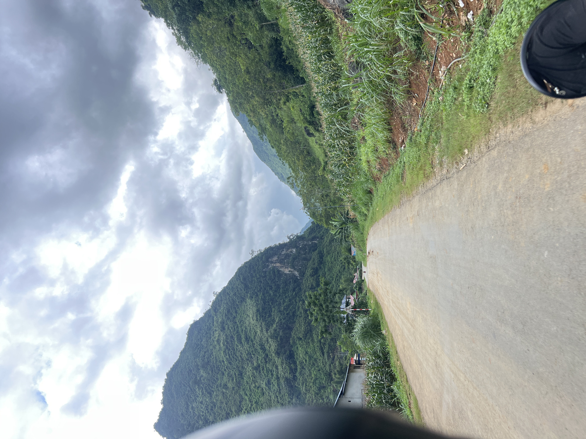 The image depicts a view of a rural road in Cán Tỷ Commune, Vietnam, characterized by steep, verdant mountains on either side. The road itself is unpaved, appearing to be a dirt or gravel track, curving gently into the distance. On the right side of the road, a field of corn stalks grows abundantly, interspersed with taller grasses and some scattered debris. The left side of the road features a few scattered buildings, including what appears to be a simple house with a white exterior, and a small agricultural field. A traffic sign is visible near the buildings. The sky above is overcast with dramatic, dark grey clouds, suggesting impending rain or a storm. The perspective suggests the photo was taken from a moving vehicle, possibly a motorcycle, given the partial view of a black helmet in the foreground. A solitary, small figure, presumably a person, is visible walking on the road in the distance, adding a sense of scale to the vast mountainous landscape. The overall impression is one of a remote, agricultural community nestled within a dramatic, natural environment.