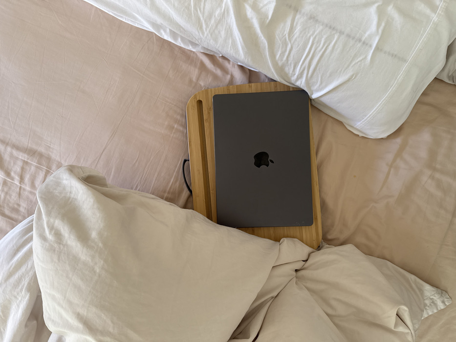 The scene depicts an interior view of a bed located in the city of Los Angeles, United States of America. The bed is covered with wrinkled light pink sheets. A rumpled light beige duvet occupies the left portion of the bed, partially overlapping a light-colored wooden or bamboo bed tray. A closed, dark gray Apple MacBook, displaying the Apple logo on its lid, rests horizontally on the bed tray. The bed tray includes a handle cut-out and a small groove on its upper surface. A rumpled white pillow is visible on the right side of the bed. No persons are present, and no active interactions are depicted.
