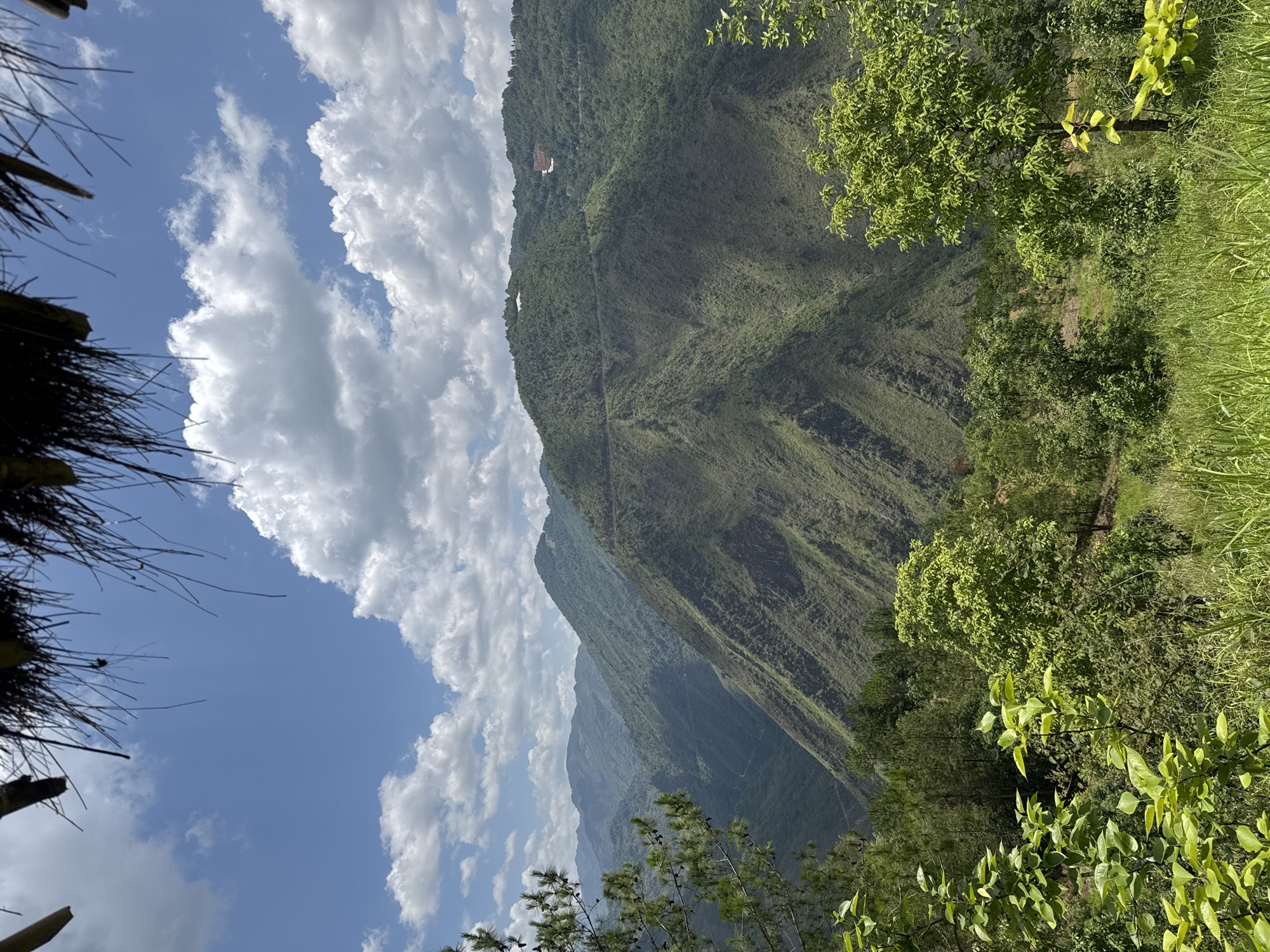 The image shows a view from Lũng Cú Commune, Vietnam, featuring a steep, green, mountainous landscape under a partially cloudy blue sky. The perspective is from within a structure with a thatched roof visible in the left corner. Lush vegetation fills the right side of the frame. A faint path or road is visible tracing along the face of the mountain. The lighting suggests a sunny day. A building can be seen near the top of the mountain.