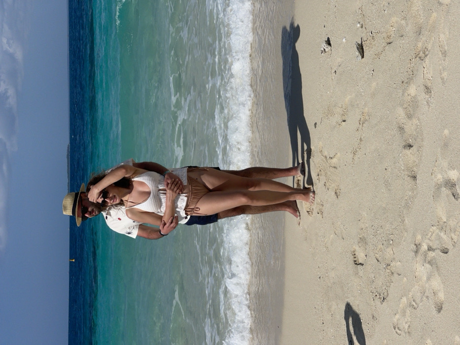 Two individuals, an adult male and an adult female, stand embraced on a light-colored sandy beach in Zanzibar, Tanzania. The male, positioned behind the female, has a beard, wears sunglasses, a straw fedora, a white short-sleeved patterned shirt, and dark shorts. His arms encircle the female's waist. The female, facing slightly left, wears sunglasses, a white crocheted-style spaghetti strap top, and light brown bikini bottoms; her right hand is raised to her forehead. Both subjects are barefoot, standing at the edge of the incoming tide where small white waves break. The ocean exhibits turquoise hues near the shore transitioning to darker blue further out. A yellow buoy is visible in the mid-ground water, with a distant landmass on the horizon under a mostly clear blue sky with sparse white clouds. Strong shadows of the subjects are cast on the sand to their right. The sandy foreground contains visible footprints and small white organic debris.