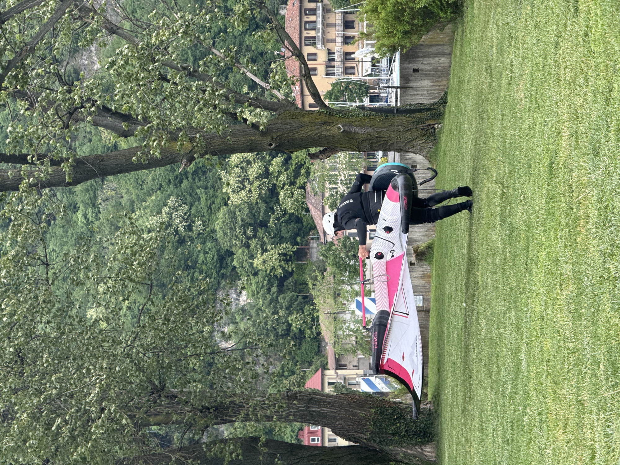 The image depicts a person on a verdant green lawn beside a body of water, likely Lake Como in Valmadrera, Italy. The individual, wearing a black wetsuit and a white helmet, is interacting with a white and pink wingfoil wing or windsurf sail, which is partially laid on the grass. Visible text on the equipment includes "CIOCC" and "GONG."
The scene suggests preparation for, or conclusion of, a water sport activity. In the background, across a concrete wall, are several buildings exhibiting typical Italian architecture with yellow-orange facades and red tiled roofs. Boats are docked nearby, indicating a small marina or harbor. Lush green trees with abundant foliage frame the foreground and mid-ground, contributing to the tranquil setting. Beyond the buildings, dense green hills and mountains rise, characteristic of the Lake Como region.
The time of day is daytime, under what appears to be soft or overcast light, as no harsh shadows are visible. The vibrant greenery suggests a mild season, such as spring or summer. The overall atmosphere is one of serene recreation in a picturesque lakeside environment.
