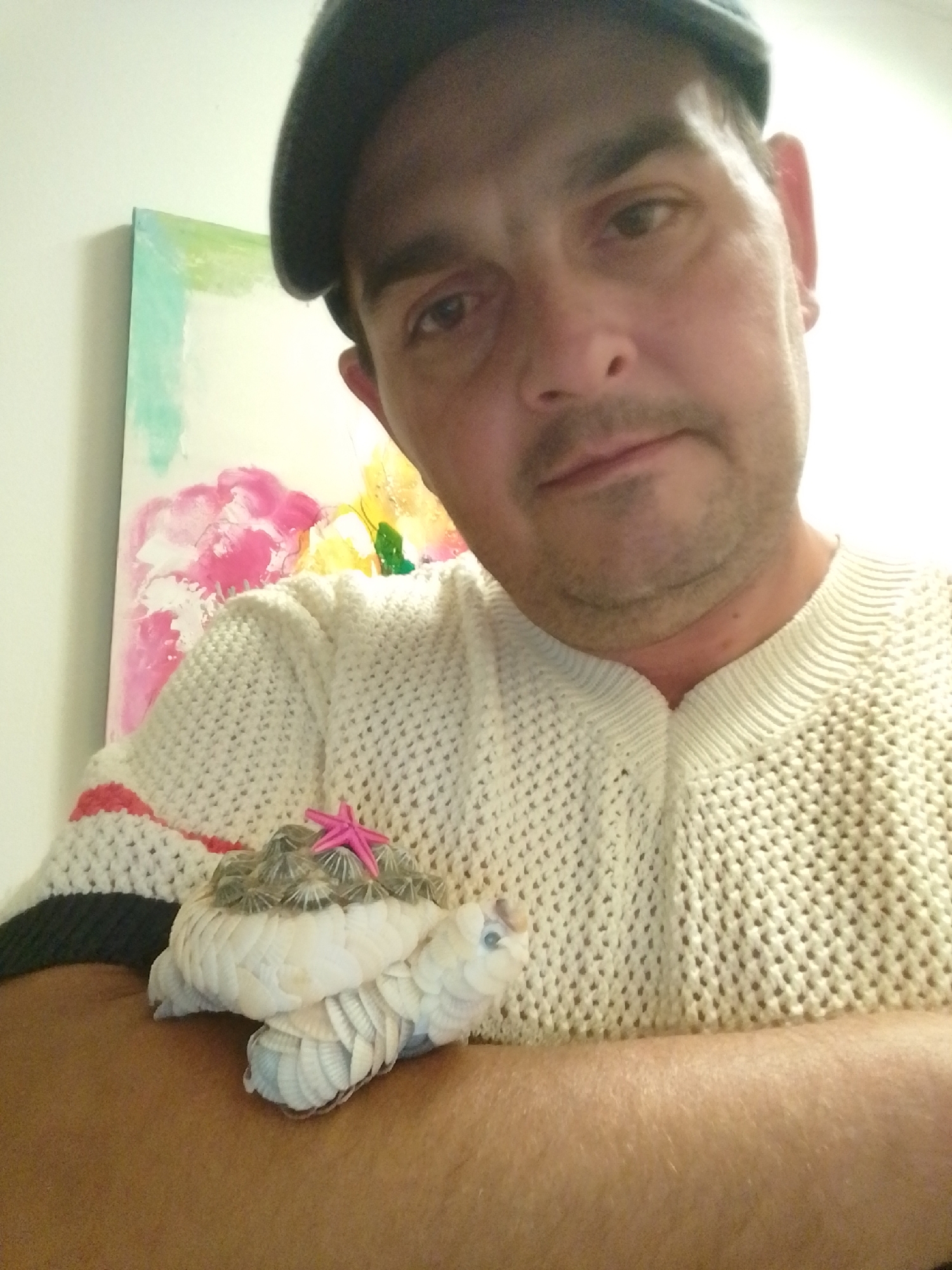A man wearing a dark beret and a cream-colored, cable-knit sweater is holding a decorative object made of shells. The object, resembling a small, plump bird or creature, has a textured top covered in grey, cap-like shells and a pink star decoration. The lower portion is formed from white shells arranged in overlapping, scale-like patterns. The man's right arm is visible, resting on his lap or a surface, with the object placed on his forearm. In the background, a portion of a painting with abstract brushstrokes of pink, yellow, and green is visible against a plain wall. The scene is lit from the front, creating a bright, diffused light. The image was captured indoors, likely a residential setting, and is a close-up shot focusing on the man and the shell creation. The overall setting is domestic, and there is no indication of the city of Wedel, Germany.