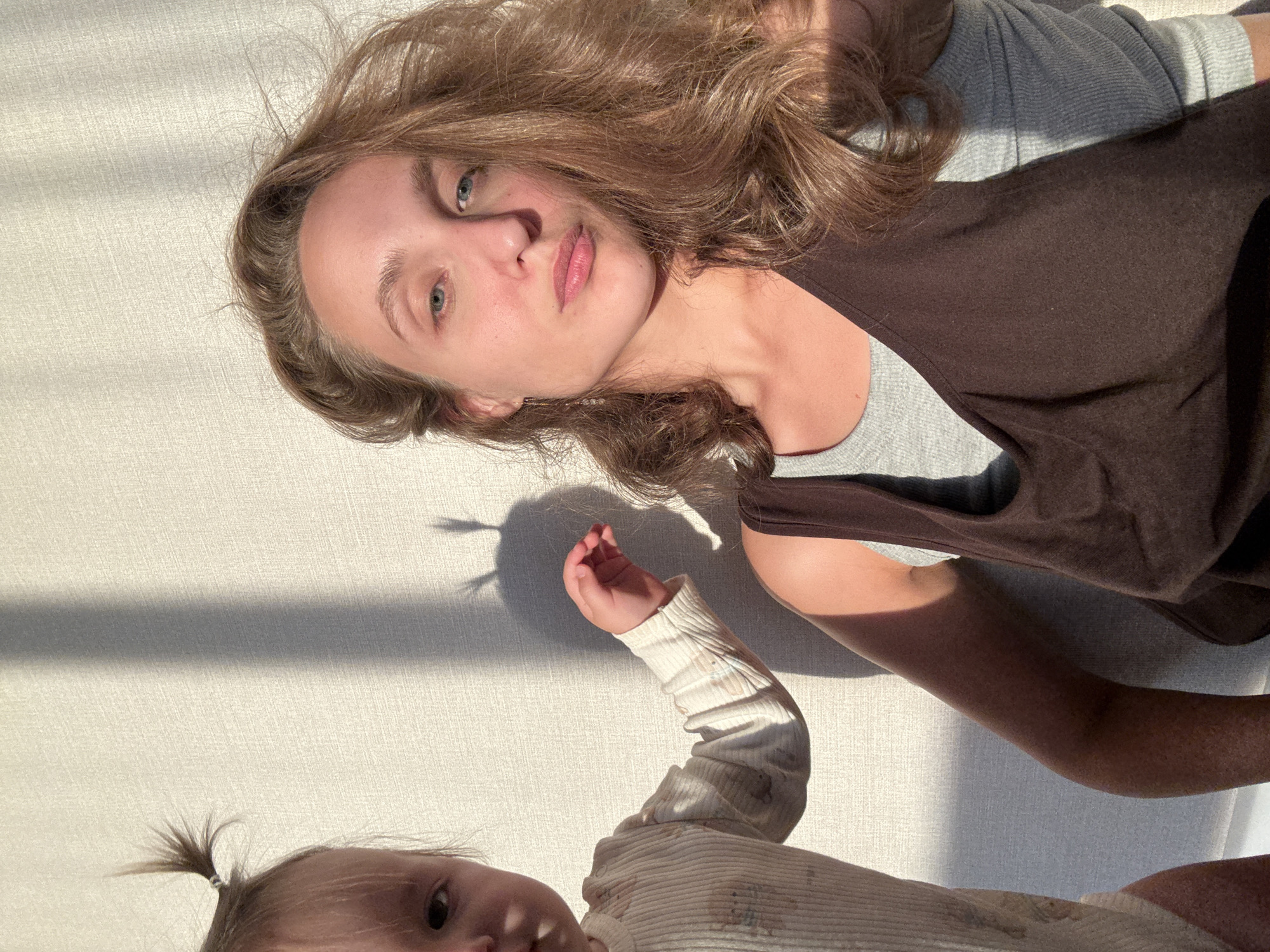 A young child with two small pigtails is partially visible on the left side of the frame, wearing a light-colored ribbed onesie with a pattern of animals. The child's left arm is extended towards the right. To the right of the child, a young woman is captured in a selfie, looking directly at the camera with a neutral expression. She has long, wavy brown hair and is wearing a grey long-sleeved shirt under a dark brown tank top. Both subjects are positioned in front of a textured, light-colored wall that casts strong shadows from an unseen light source, creating vertical stripes across the scene. The lighting suggests late afternoon or early morning sun. The setting appears to be indoors. There is no discernible indication of the city of Подшивалово, Russia, within the image itself.