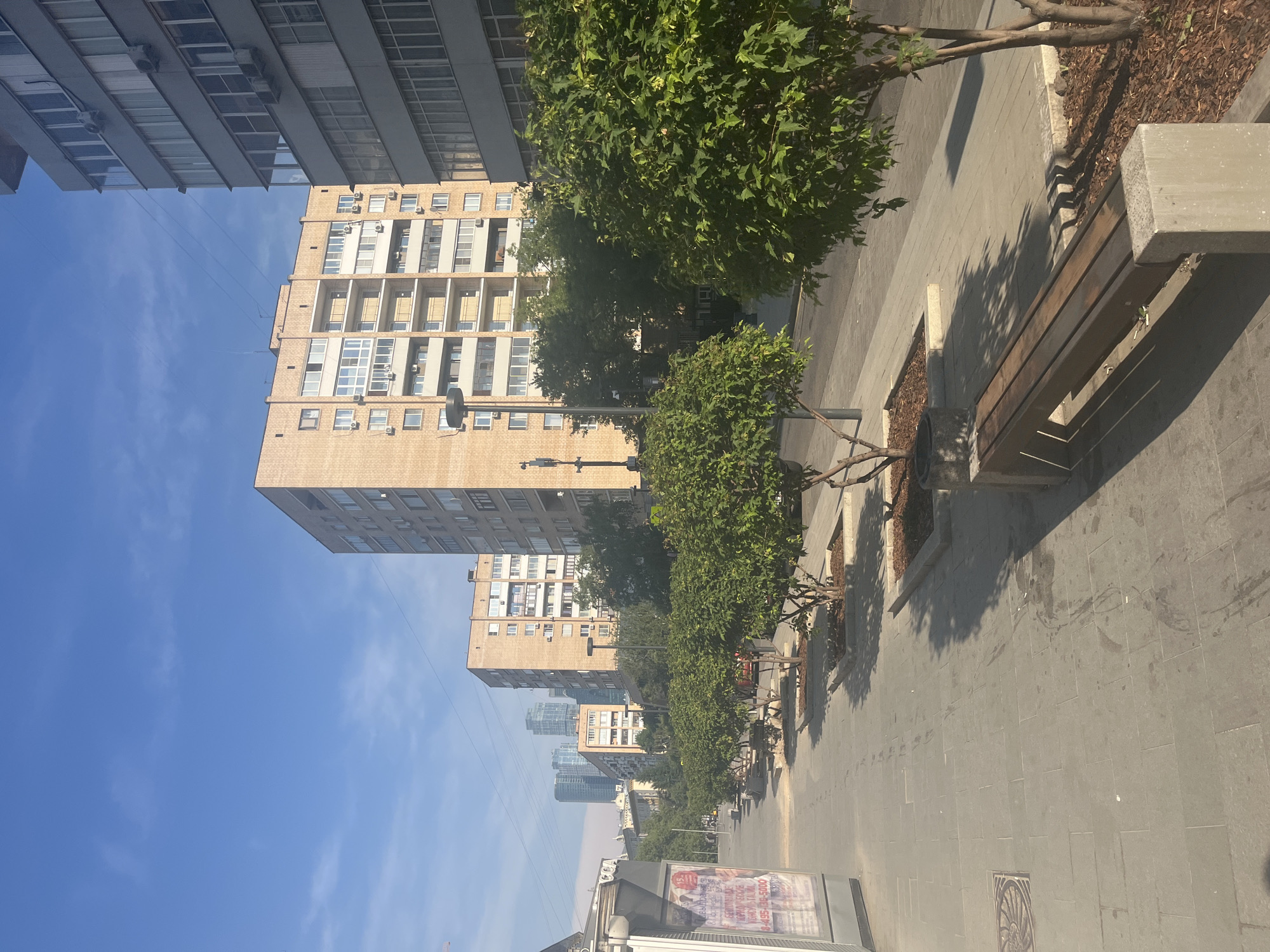 The image depicts a sunny day on a wide, paved pedestrian walkway in Moscow, Russia. The walkway is lined with several small, young trees planted in raised, square beds filled with wood chips. A wooden bench with concrete supports is situated on the right side of the walkway, casting a distinct shadow.
In the background, several multi-story apartment buildings are visible. The closest building on the right is a tall, grey structure with numerous windows and balconies. To its left, a lighter-colored, brick apartment building with a more traditional design stands. Further down the street, more apartment buildings and some modern skyscrapers can be seen in the distance. The sky is a clear blue with a few wispy clouds.
On the far left of the image, a transparent structure with advertisements visible inside is present, possibly a bus stop shelter or kiosk. The ground is paved with large, rectangular grey tiles, and there are some dark stains and footprints visible on the walkway. A manhole cover with an ornamental design is also present on the left. The overall impression is an urban environment with a focus on pedestrian space and greenery.