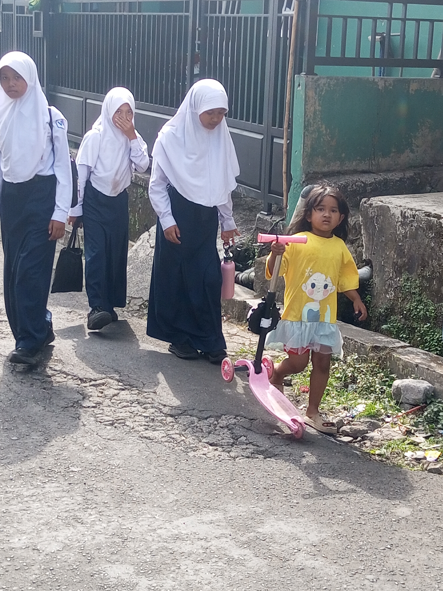 The image depicts a street scene in Krasak, Indonesia, on a sunny day. Three young females, dressed in school uniforms consisting of white hijabs and long navy skirts, are walking from left to right across a cracked asphalt road. The first girl on the left, partially obscured, wears a white shirt and a navy skirt. The second girl in the center covers her mouth with her hand, wearing a white shirt and navy skirt, and carries a black tote bag. The third girl on the right also wears a white shirt and navy skirt and holds a pink water bottle. To the right of the schoolgirls, a younger female child, with long dark hair, stands facing forward. She is wearing a yellow t-shirt with a cartoon graphic and a light blue ruffled skirt. The child is holding the handlebars of a pink scooter, which is positioned in front of her on the road. The background features a dark metal fence, a green wall, and a concrete structure with some overgrown vegetation and scattered rocks. The lighting suggests it is daytime, with shadows cast on the road.