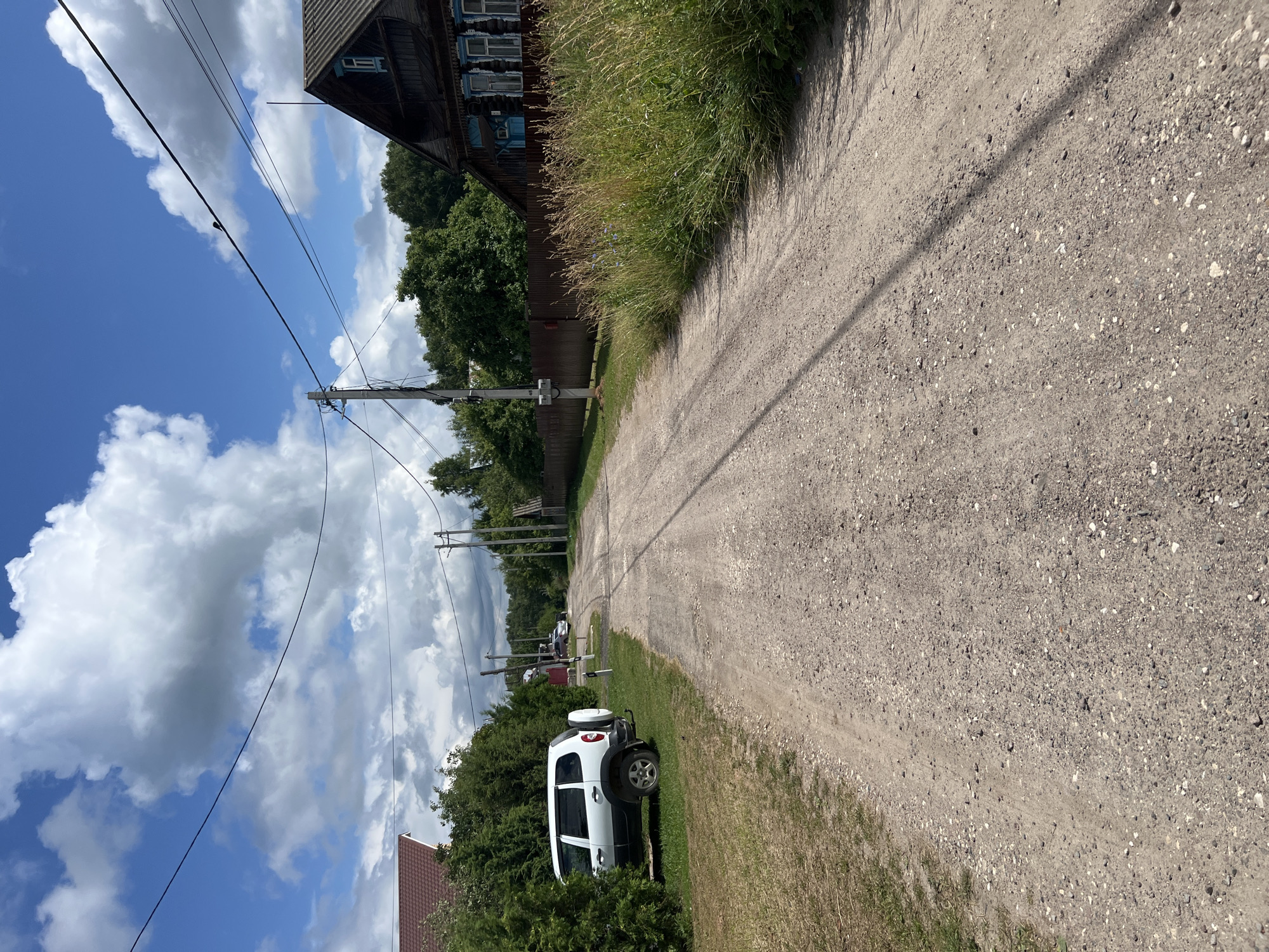 A dirt road winds through a rural area in Vyazniki, Russia, flanked by dense green foliage and wooden structures. On the left, a white SUV is parked amidst tall evergreen bushes. Further down the road, a person can be seen near a wooden fence. To the right, a weathered, dark wooden house with blue window frames stands out, bordered by tall, overgrown grass. Power lines stretch across the blue sky, which is dotted with white, fluffy clouds. The road itself is a mix of dirt and gravel, with tire tracks visible.
