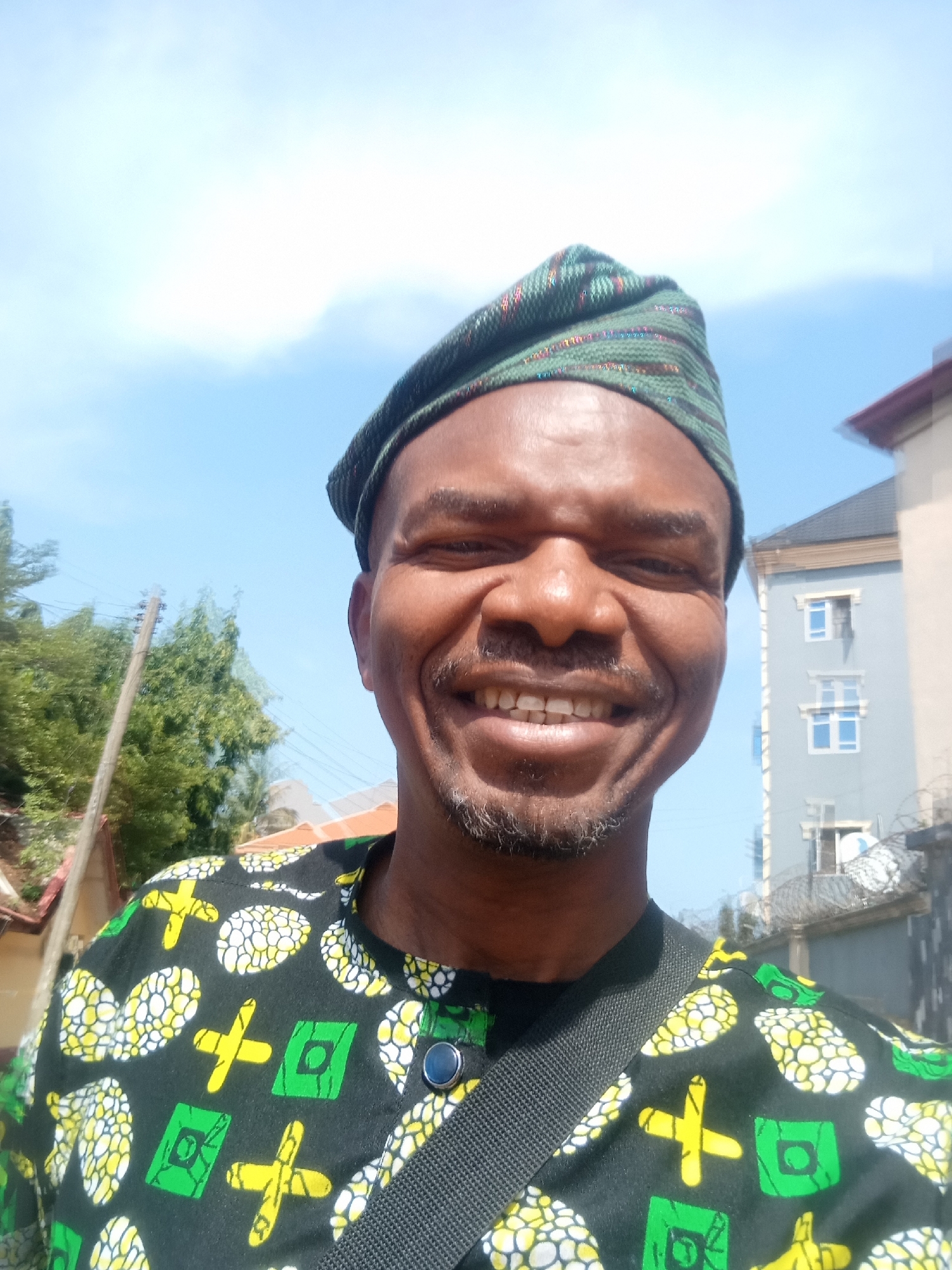 A smiling man wearing a traditional patterned shirt and a green cap is the subject of the photograph. The shirt is predominantly black with patterns of white, yellow, and green. He is also wearing a black strap across his chest, likely for a bag or camera.
Behind the man, there is a building with several windows and what appears to be barbed wire on top of a wall. There are also trees and utility poles visible in the background. The sky is a clear blue. The image likely originates from Aba, Nigeria, as requested. No specific location details are discernible.