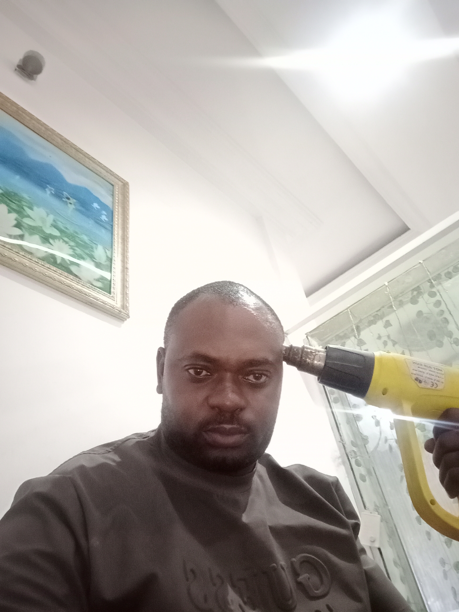 A man is holding a yellow heat gun pointed towards his head. The scene appears to be indoors, possibly inside a home, as indicated by the walls, ceiling, painting, and patterned curtains. The location is identified as Gwarinpa, Nigeria. The man is dark-skinned, with a shaved head, and wearing a gray shirt.