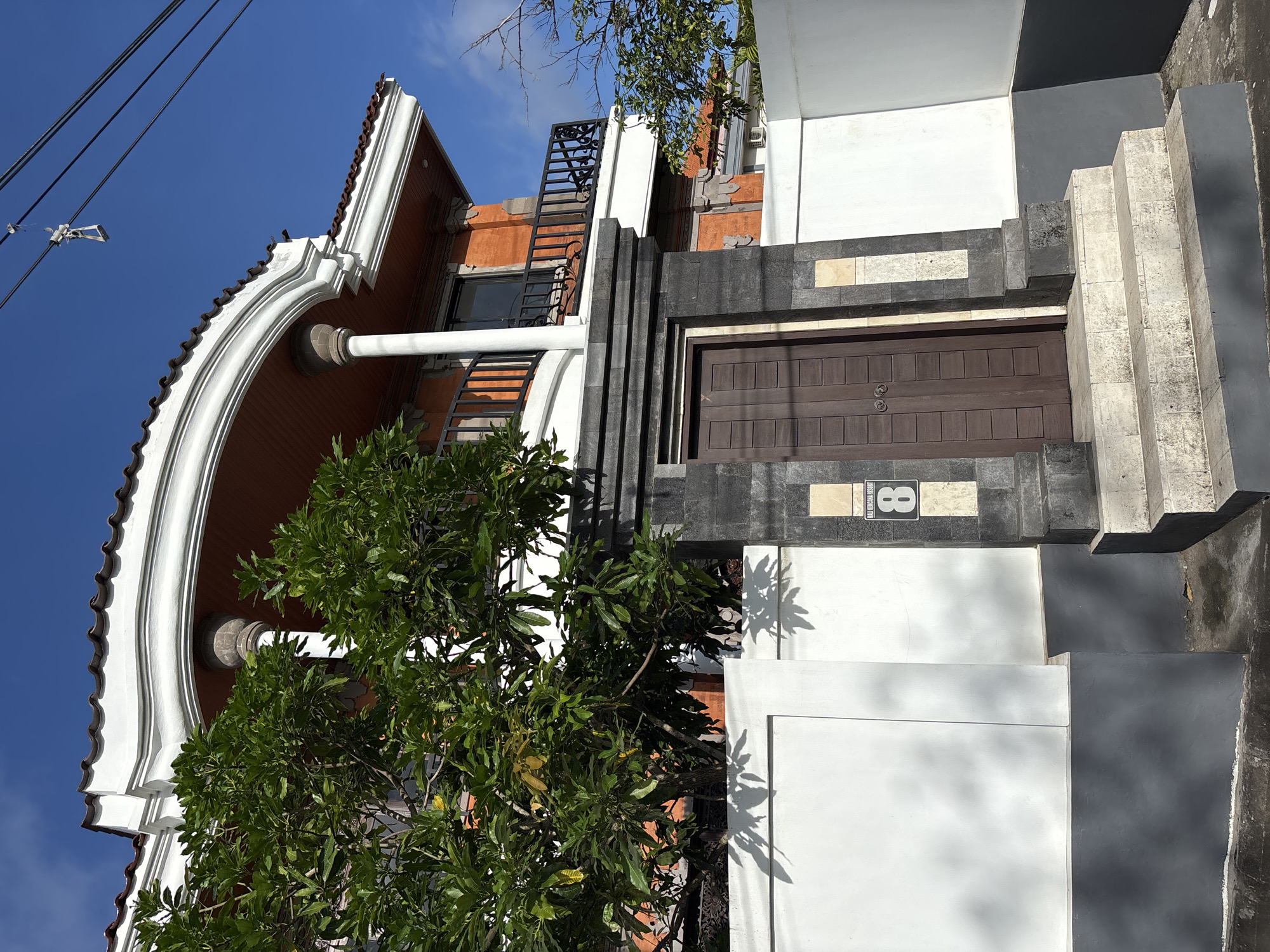 The photograph shows a two-story building with a distinctive architectural style, located in Kuta Selatan, Indonesia. The facade is a mix of white, gray, orange and brown colors and features an arched roofline, a balcony, and a ground-floor entryway, with a ladder-like structure visible. The entryway is centered and composed of dark stone and a wooden door with a sign displaying "8". Green foliage obscures part of the building's facade. Electrical wires cross above the structure against a clear blue sky. A stairway ascends to the door. The photograph appears to have been taken from a slightly low angle.