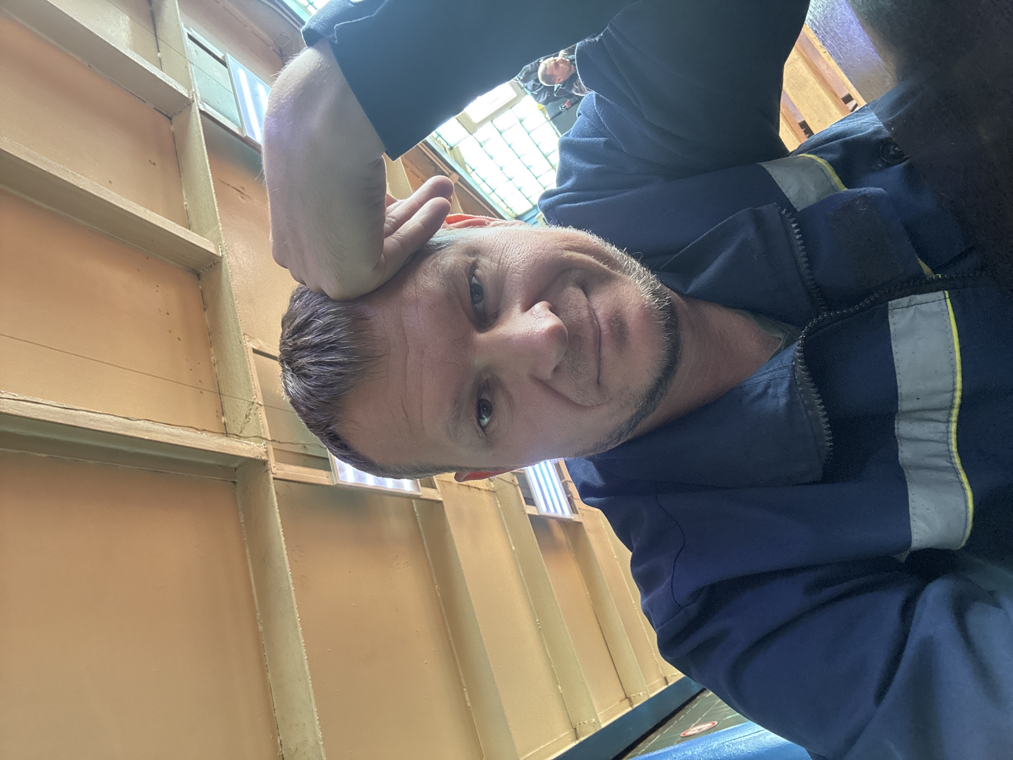 The image is a selfie taken by a man indoors. He is wearing a dark blue jacket with a reflective stripe across the chest. His right hand is on his forehead. The man has short, light brown hair and stubble. In the background, there is a ceiling made of wooden beams and panels, and a window with panes. There are other people visible in the background, out of focus. One person appears to be a man wearing a dark jacket and another person is partially visible. The scene appears to be a public indoor space, possibly a waiting area or a covered walkway. The specific location in Oskemen, Kazakhstan is not determinable from the visual cues.