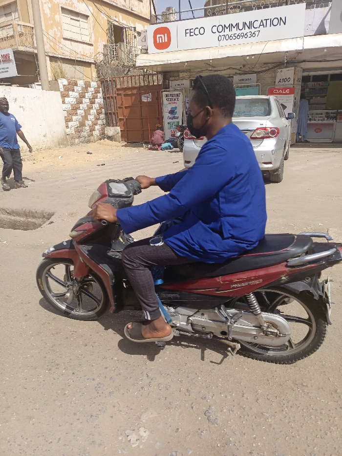 The image presents a street scene in Fagge A, Nigeria, under bright daylight. In the foreground, a man with dark skin is seated on a red and black motorcycle, holding the handlebars. He wears a blue long-sleeved tunic, dark patterned trousers, brown slides, a black face mask, and sunglasses perched on his head. The motorcycle is identified as a 'PASSPORT' model, featuring silver-colored engine components and spoked wheels.
To the immediate left of the central figure, a second man, wearing a blue t-shirt and dark pants, walks towards the left, with his left foot raised. Behind him, a light-colored building with a section of brown and white checkered wall is visible, alongside a rusted metal gate topped with barbed wire.
In the background, a commercial storefront is present. A prominent white sign above it displays a red 'Mi' logo, the text "FECO COMMUNICATION," and a phone number "0706569367." The storefront features product displays, including signs for "SAMSUNG," "A100," and "OALE." A silver sedan is parked in front of this store, partially obscuring the entrance. Near the storefront entrance, a person dressed in red/pink clothing is seated on the ground. Another sign advertising "COMPUTERS SERVICE" and "EasyBuy" is also visible.
The ground surface is an uneven, light-colored, dusty asphalt or dirt road.