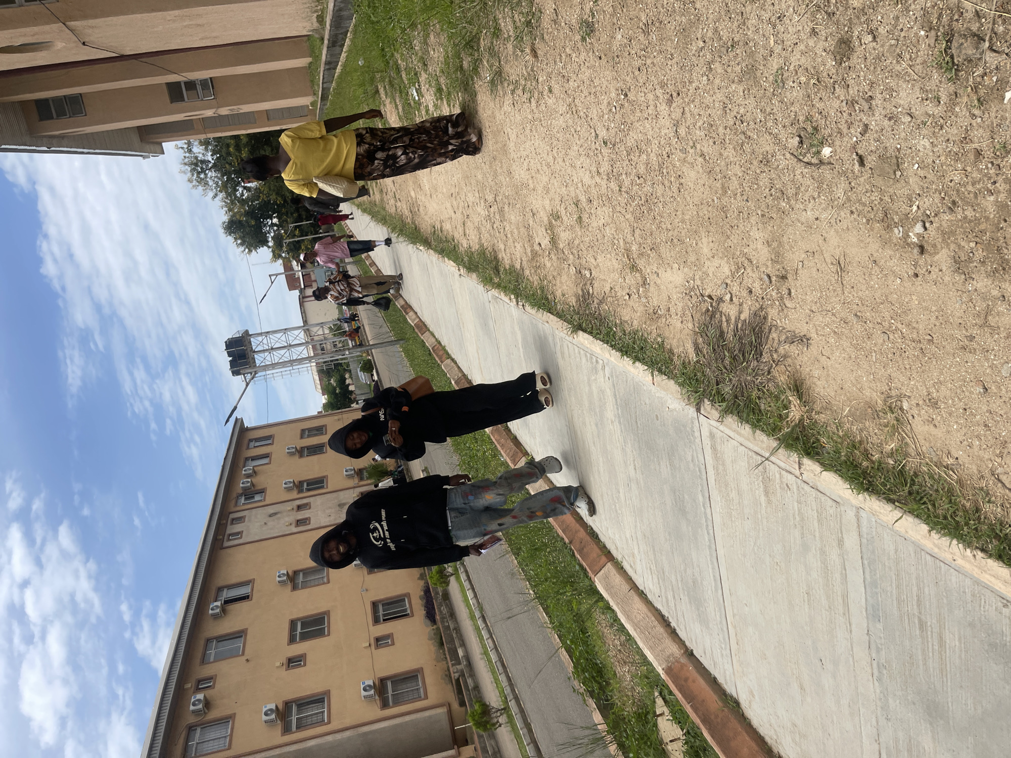 The image depicts an outdoor scene in the city of Jos, Nigeria, under a cloudy sky. In the foreground, two individuals, a man and a woman, walk side-by-side on a concrete sidewalk. The man is wearing a black hooded sweatshirt and blue jeans with colorful paint splatters. The woman is in a black hooded sweatshirt and black pants. Behind them, several other people are walking in the same direction on the sidewalk. Further in the background, a tan building with multiple windows and air conditioning units lines the left side of the street. On the right side of the scene, another tan building with rectangular windows is visible. A dirt path with sparse vegetation runs alongside the sidewalk. A water tower structure is visible in the distance.