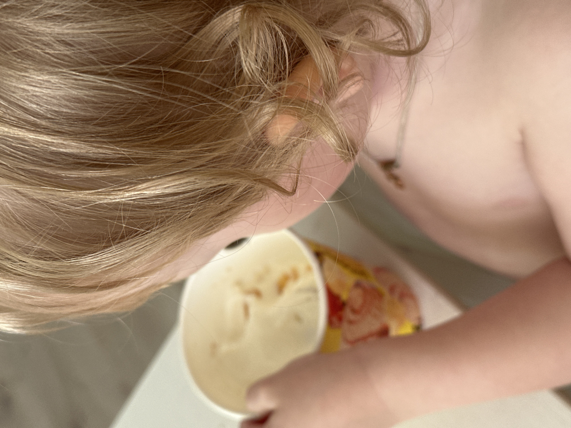 A blonde-haired child is bent over, looking into a paper cup. The cup contains a pale, creamy substance with some orange specks. The child's shoulder and arm are visible, along with a hint of a necklace. The background is a light-colored surface. It's not possible to discern the location or city without further information.