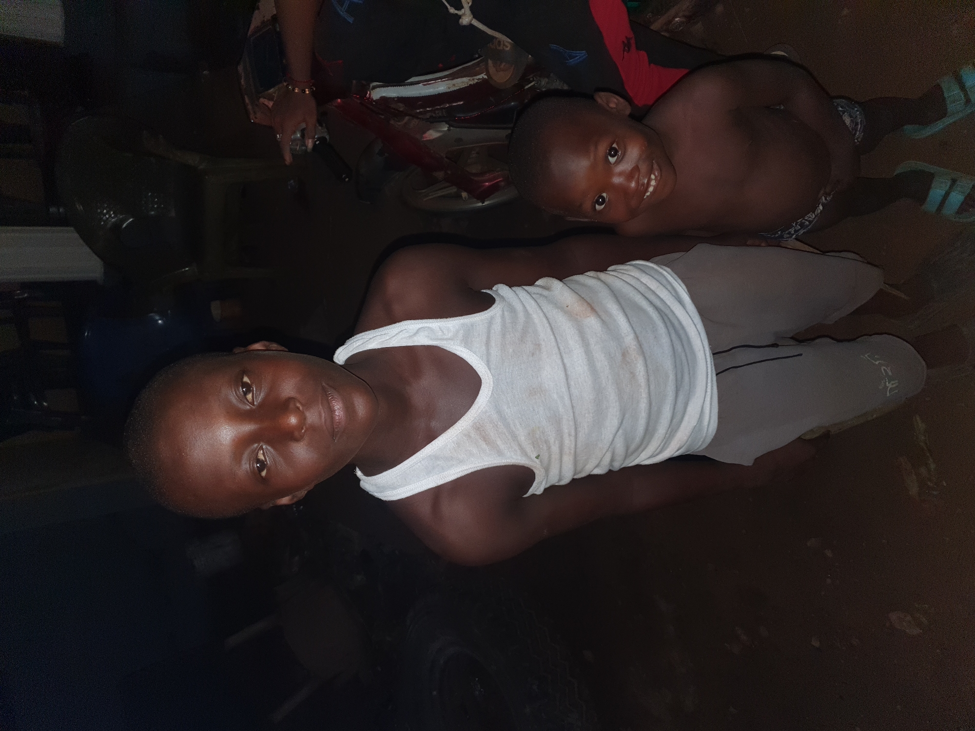 This dimly lit vertical image, possibly taken at night in Idah, Nigeria, features two young boys looking towards the camera and smiling.
In the foreground, a boy is positioned towards the left side of the frame. He has dark skin and a short haircut, looking directly at the camera with a gentle smile. He is wearing a white sleeveless tank top and light grey pants with some faint, possibly Arabic, text visible on the left leg, appearing like "لل".
Behind him, slightly to the right and appearing to be at a higher elevation, is a second boy. He also has dark skin and a broad smile, looking playfully towards the camera. This boy is bare-chested and wears patterned shorts or underwear. Light blue sandals are visible on his feet. An "adidas" logo can be seen on a slipper worn by an adult hand behind him.
Partially visible behind the second boy and the adult hand is the red and white frame of what appears to be a motorcycle or scooter. The adult's hand, adorned with a bracelet, rests on a handle of this vehicle. The background is mostly dark and indistinct, suggesting an indoor setting or a very poorly lit outdoor area. The overall mood of the image is joyful and candid, captured with direct flash in a low-light environment.