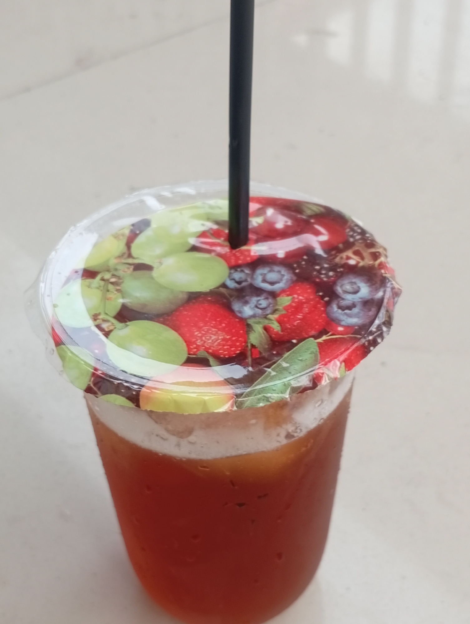 A close-up, eye-level shot captures a plastic cup filled with iced tea, placed on a textured, off-white surface. The cup is sealed with a clear plastic lid adorned with a colorful printed design featuring various berries and grapes, including strawberries, blueberries, and green grapes. A black plastic straw protrudes from the center of the lid. Condensation droplets are visible on the exterior of the cup. The background is blurred, indicating an outdoor or semi-outdoor setting with the suggestion of a soft, light-colored wall or structure. The overall impression is that of a refreshing beverage on a casual day, potentially from a local vendor in Depok, Indonesia.