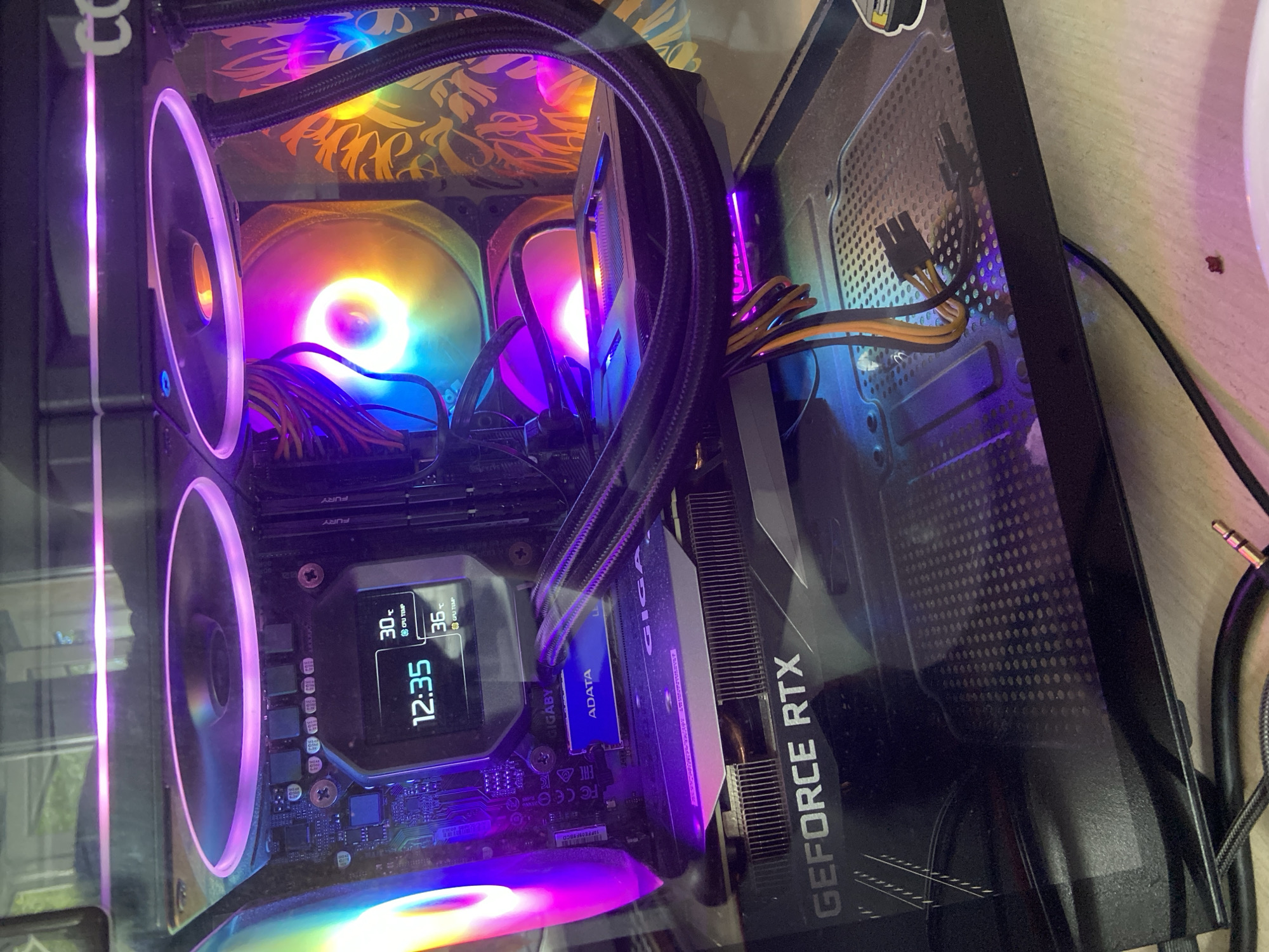 The image shows the interior of a computer tower, likely a gaming PC, with visible components illuminated by RGB lighting. A central liquid cooler with RGB fans is prominently displayed, with one fan showing a rainbow spectrum of colors and the other a pinkish-purple hue. The motherboard is visible with RAM sticks labeled "FURY" and an ADATA SSD. A graphics card with "GEFORCE RTX" branding is installed below the motherboard. A small digital display on the CPU cooler shows the time as 12:35 and CPU temperatures of 30°C and 36°C. The computer case appears to have a glass side panel, allowing for a clear view of the internal components. Cables, including power connectors, are neatly routed. The background is blurred, but appears to be an indoor setting with a wooden surface. There are no people or animals present in the image. The location of Beloretsk, Russia is not discernible from the image itself.