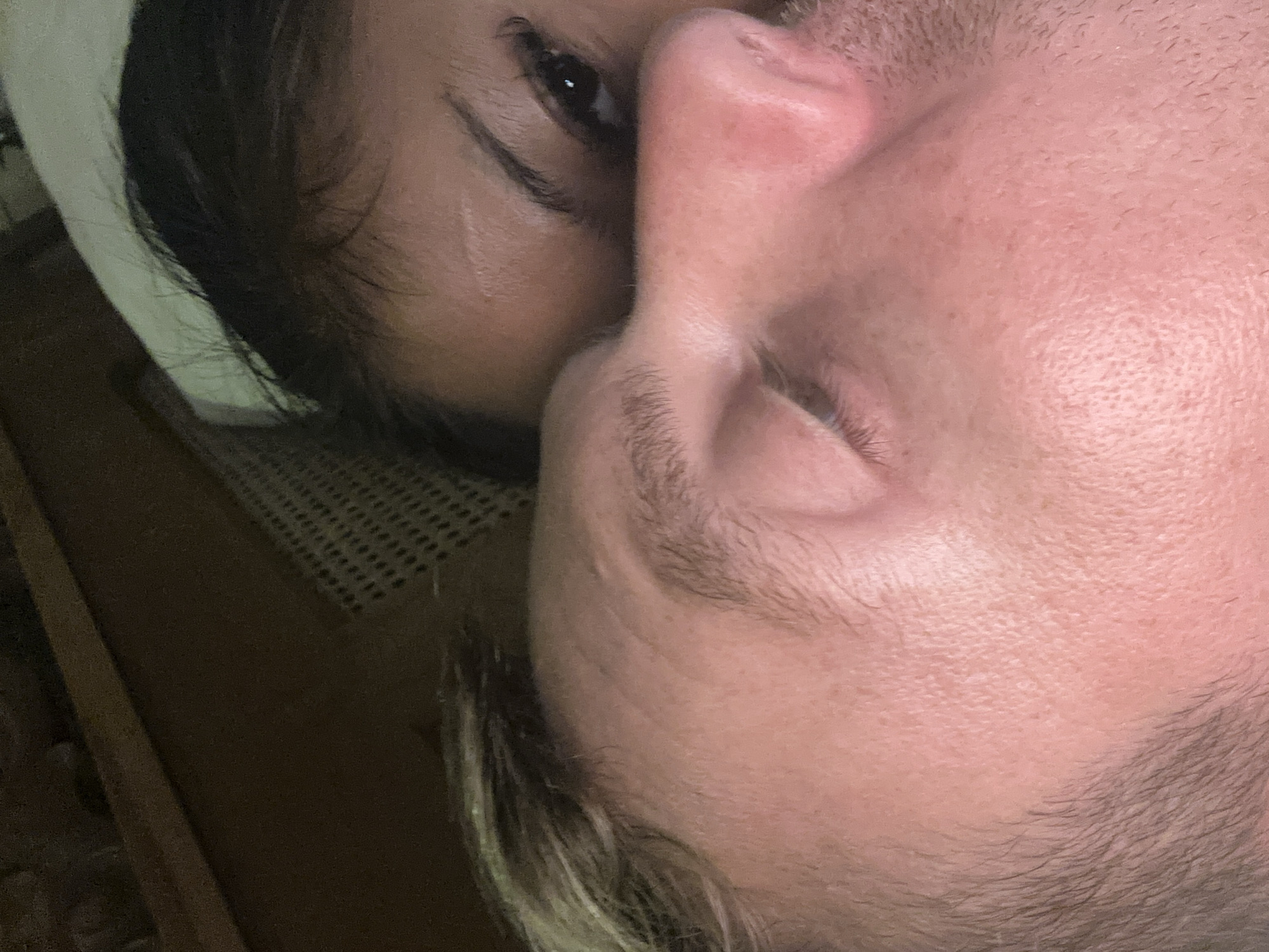 A close-up, angled view features the faces of two individuals. The person on the left has dark hair, an eye open, and a visible scar above the eyebrow. The person on the right has light skin and facial hair. The faces are close together. In the background, there is a wooden surface with a perforated pattern. The setting is unknown, but given the mention of Ubud District, Indonesia, it could potentially be a private residence or accommodation there.