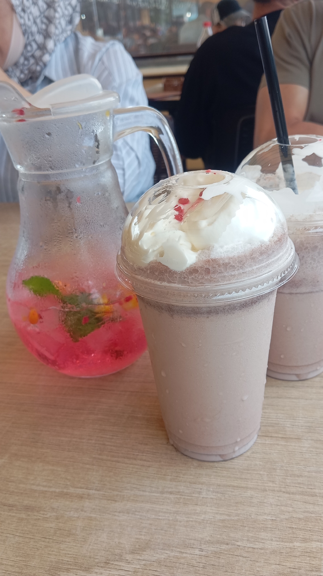 In an indoor setting, possibly a cafe or restaurant, a clear glass pitcher filled with a pink liquid, ice, mint leaves, and small red and yellow pieces sits on a light-colored wooden table. To the right of the pitcher are two clear plastic cups, each containing a blended drink topped with whipped cream and garnished with red sprinkles. A black straw is visible in the front cup. In the background, blurry figures of people can be seen, some wearing casual clothing. The overall lighting suggests a daytime scene. It is not possible to determine the specific location within Nalchik, Russia, from the provided image.